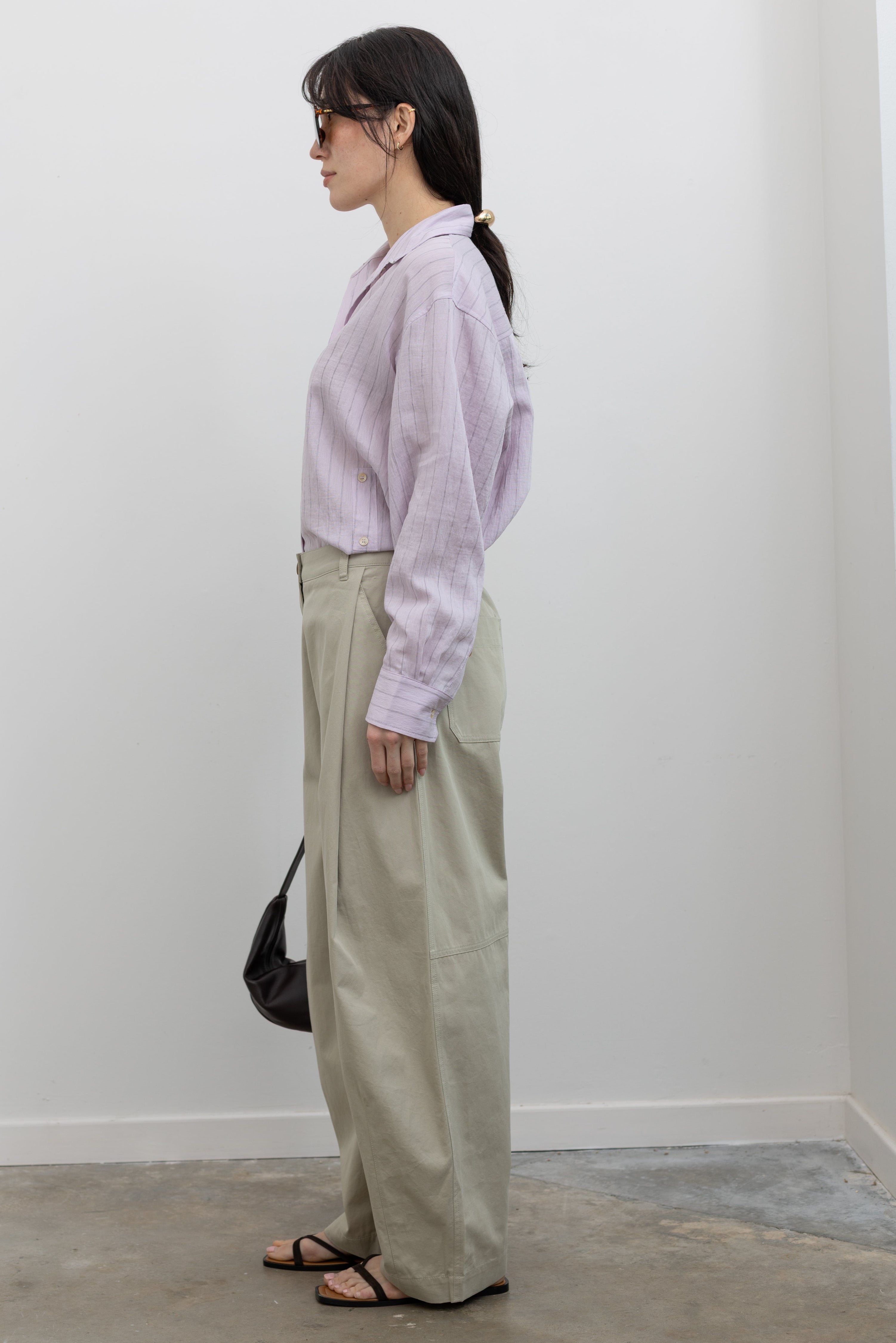 Full length side view of a woman standing wearing sophisticated spring fashion in Mijeong Park beige 100% cotton baggy barrel leg pants featuring curved outseams that create architectural volume and a partially elasticated waistband and belt loops for a tailored look paired with an elevated classic Mijeong Park pink stripe button down shirt with a St. Agni leather bag and strappy sandals at an Alas Eius store. 