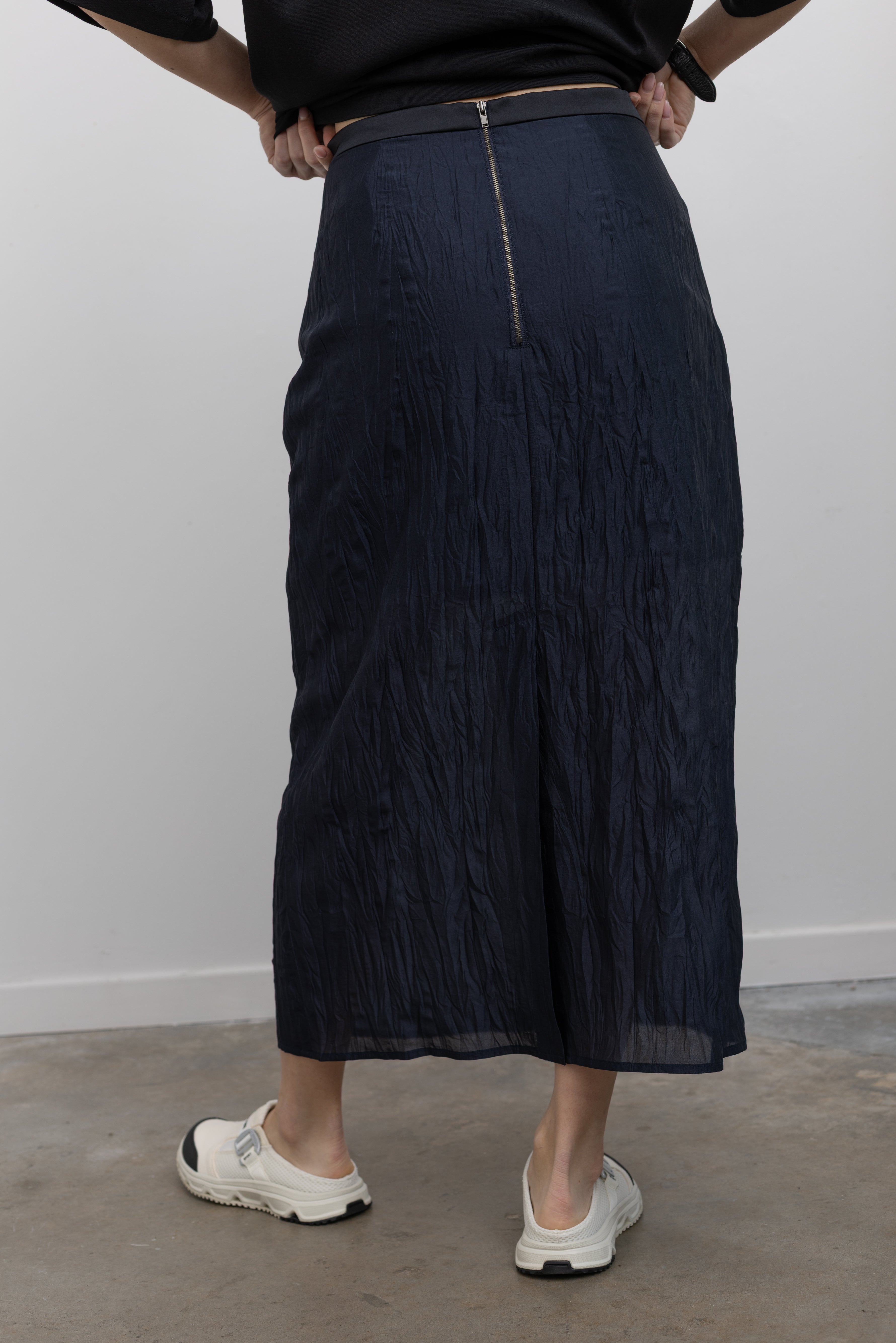 Close-up back view of a woman standing wearing contemporary spring fashion in a Mijeong Park navy blue lightweight, semi-sheer crinkled fabric lined midi skirt paired with a Mijeong Park black oversized t-shirt and slide Salomon sneakers at an Alas Eius store. 