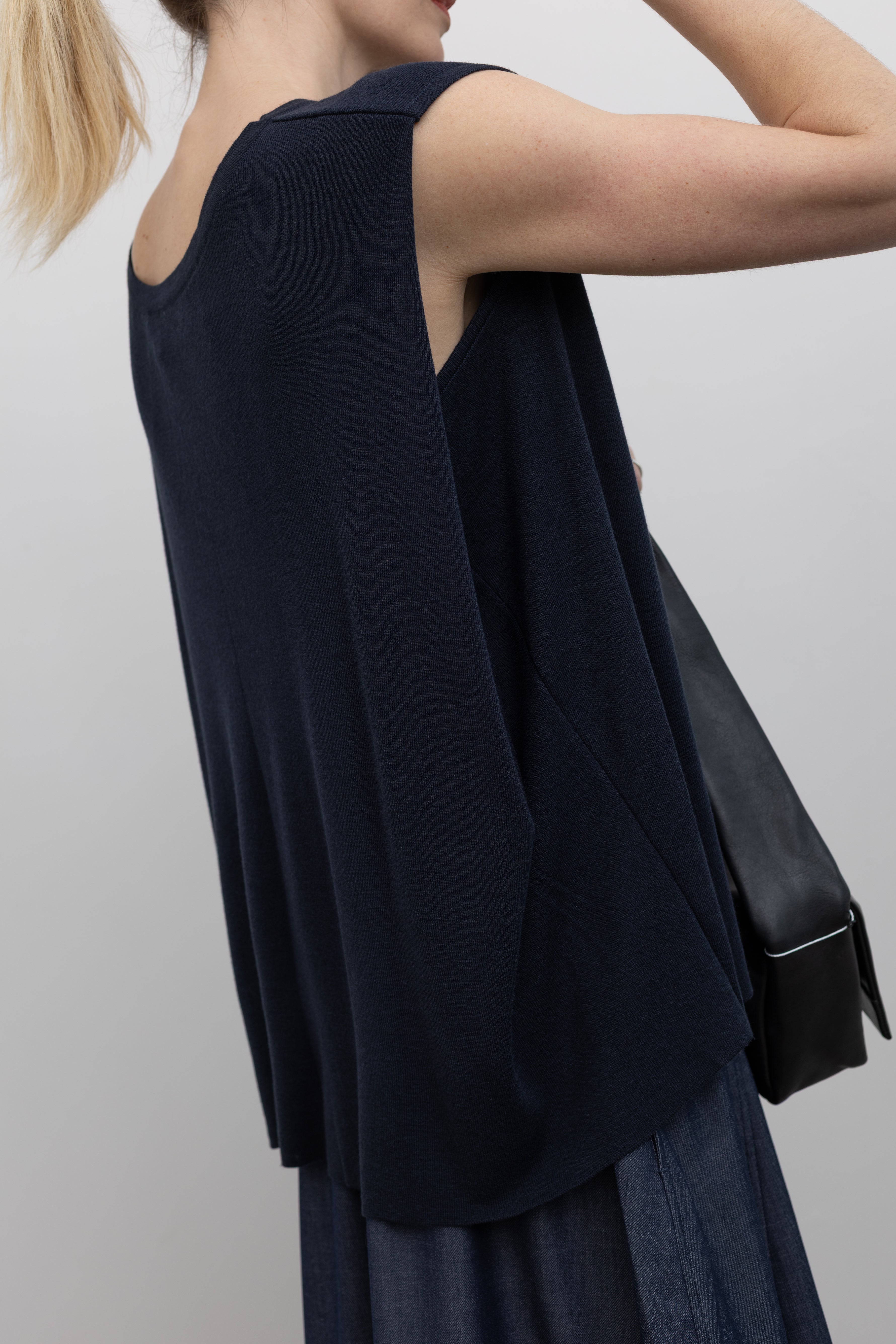 Close-up detailed view of a woman wearing a relaxed fit Mijeong Park navy blue sleeveless top with a draped back and a curved front hem at an Alas Eius store. 