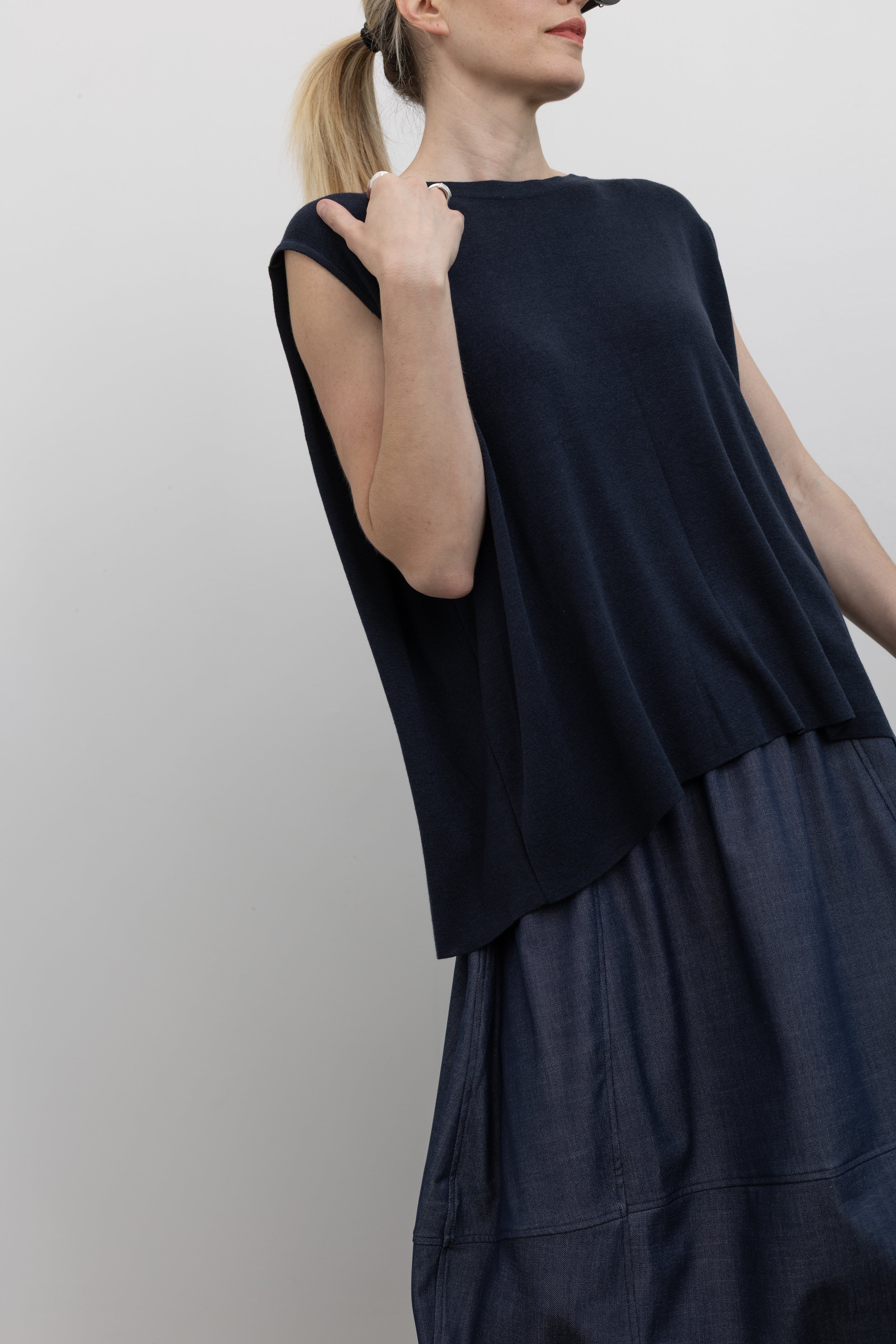 Close-up 3/4 front view of a woman wearing a relaxed fit Mijeong Park navy blue sleeveless top with a draped back and a curved front hem at an Alas Eius store. 