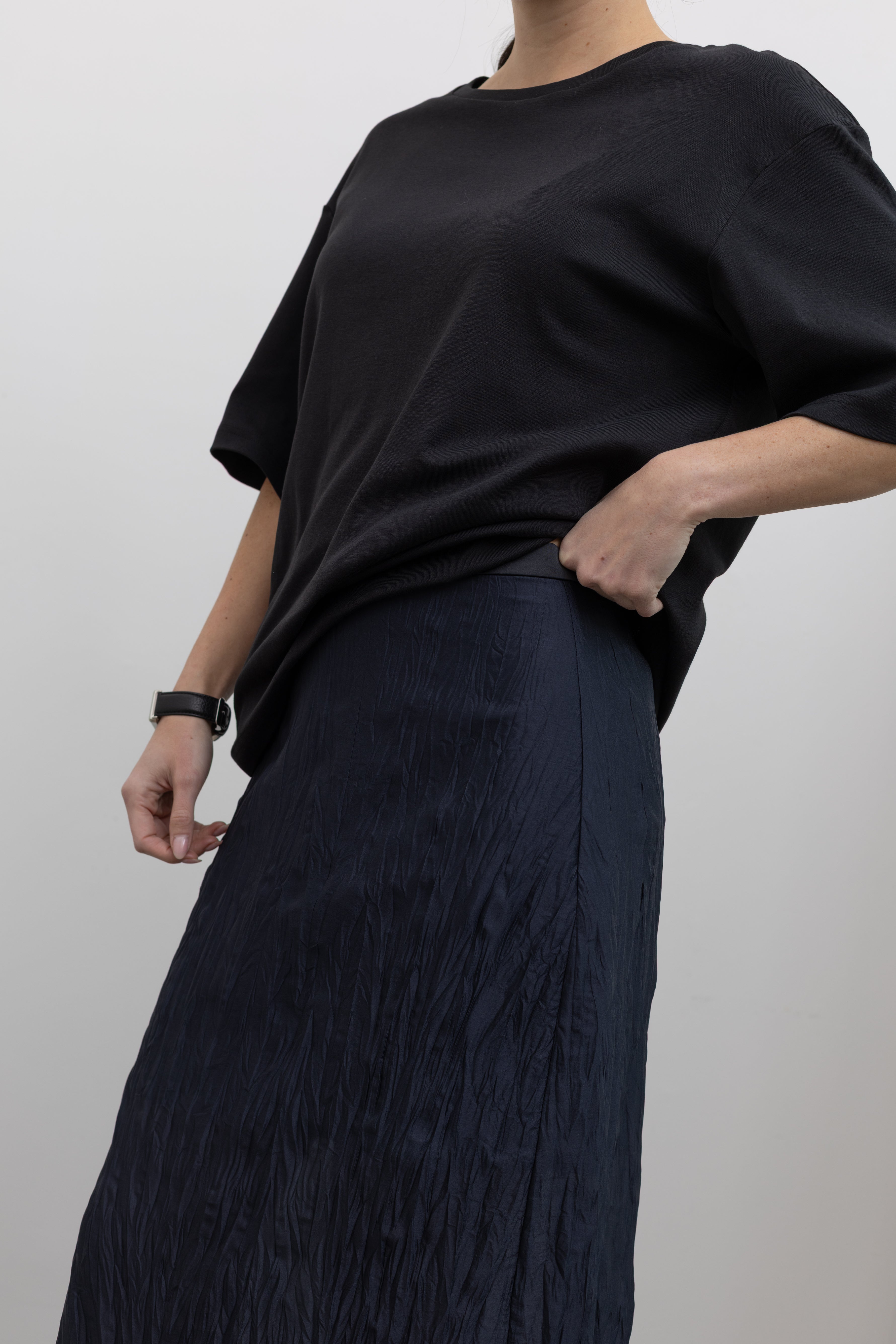 Close-up back view of a woman standing wearing flattering spring fashion in a Mijeong Park navy blue lightweight, semi-sheer crinkle fabric lined midi skirt paired with a Mijeong Park black oversized t-shirt at an Alas Eius store. 