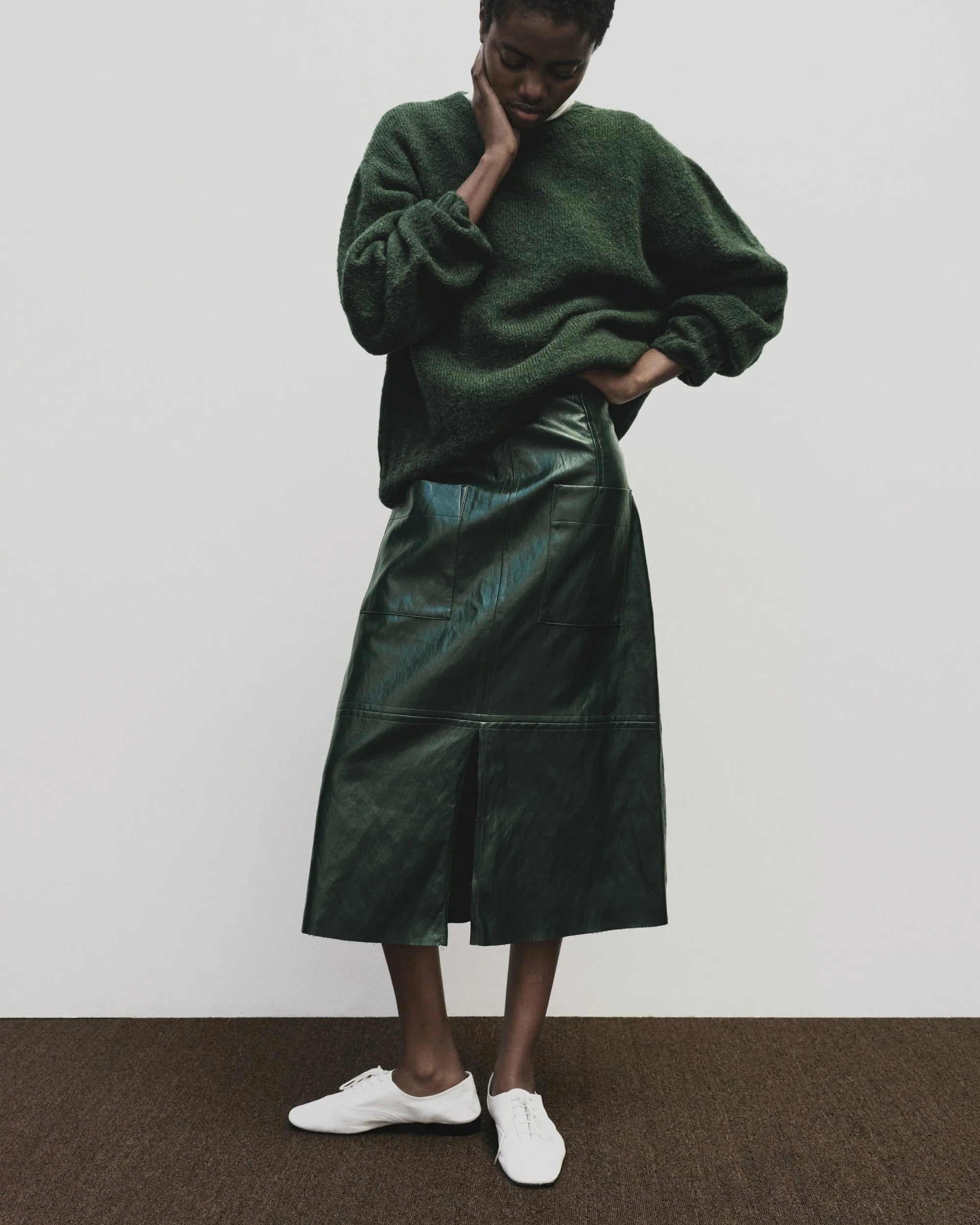 Person wearing a green sweater and leather skirt with white shoes on a plain background