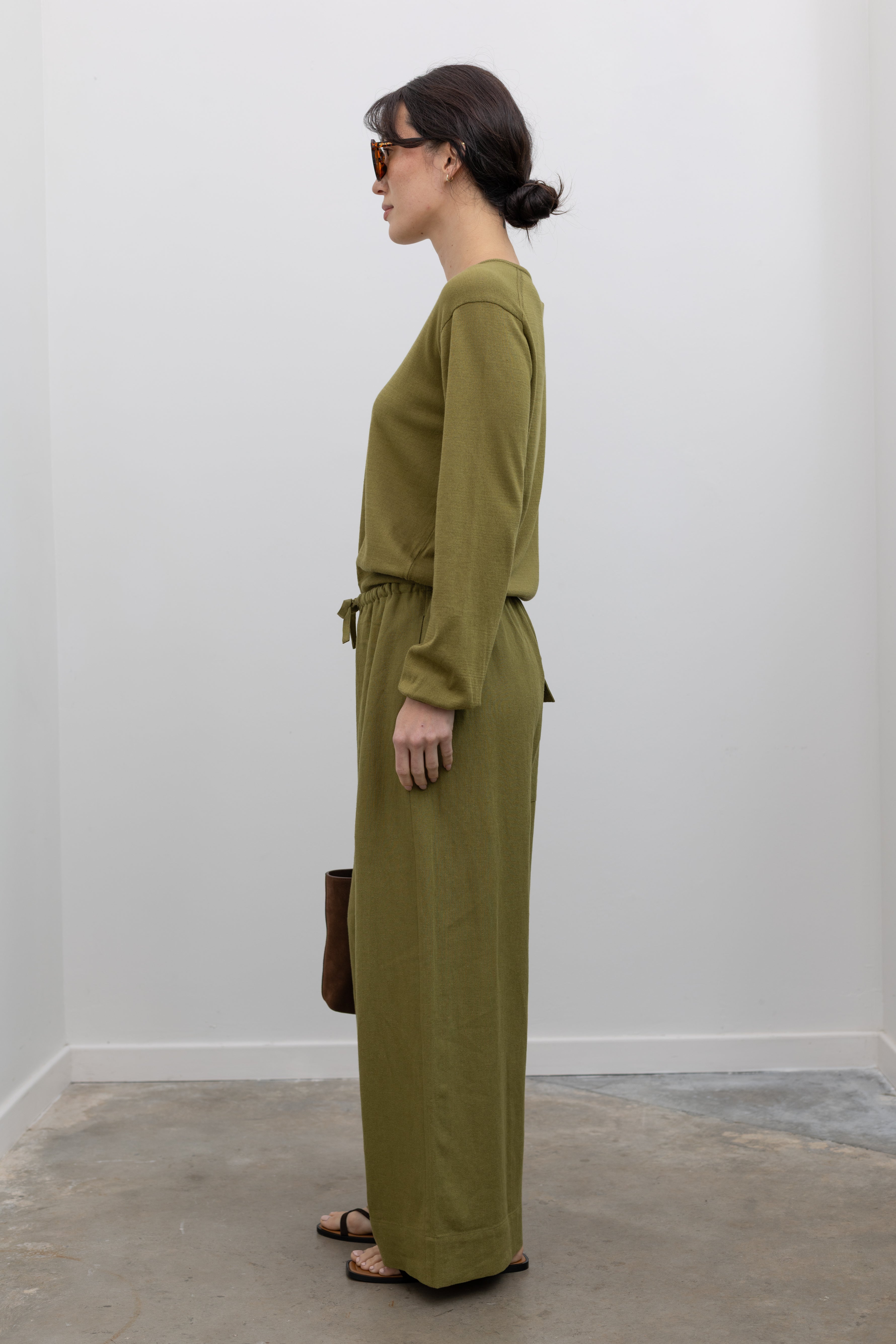 Full length side view of a woman standing wearing elevated casual spring fashion in the Mijeong Park Green Cotton Blend V-Neck Knit Top paired with Mijeong Park Green Linen Blend Wide Leg Cropped Pants featuring a comfortable drawstring waist, front and back patch pockets, and a cropped length suitable for a minimalist, versatile wardrobe at an Alas Eius store. 