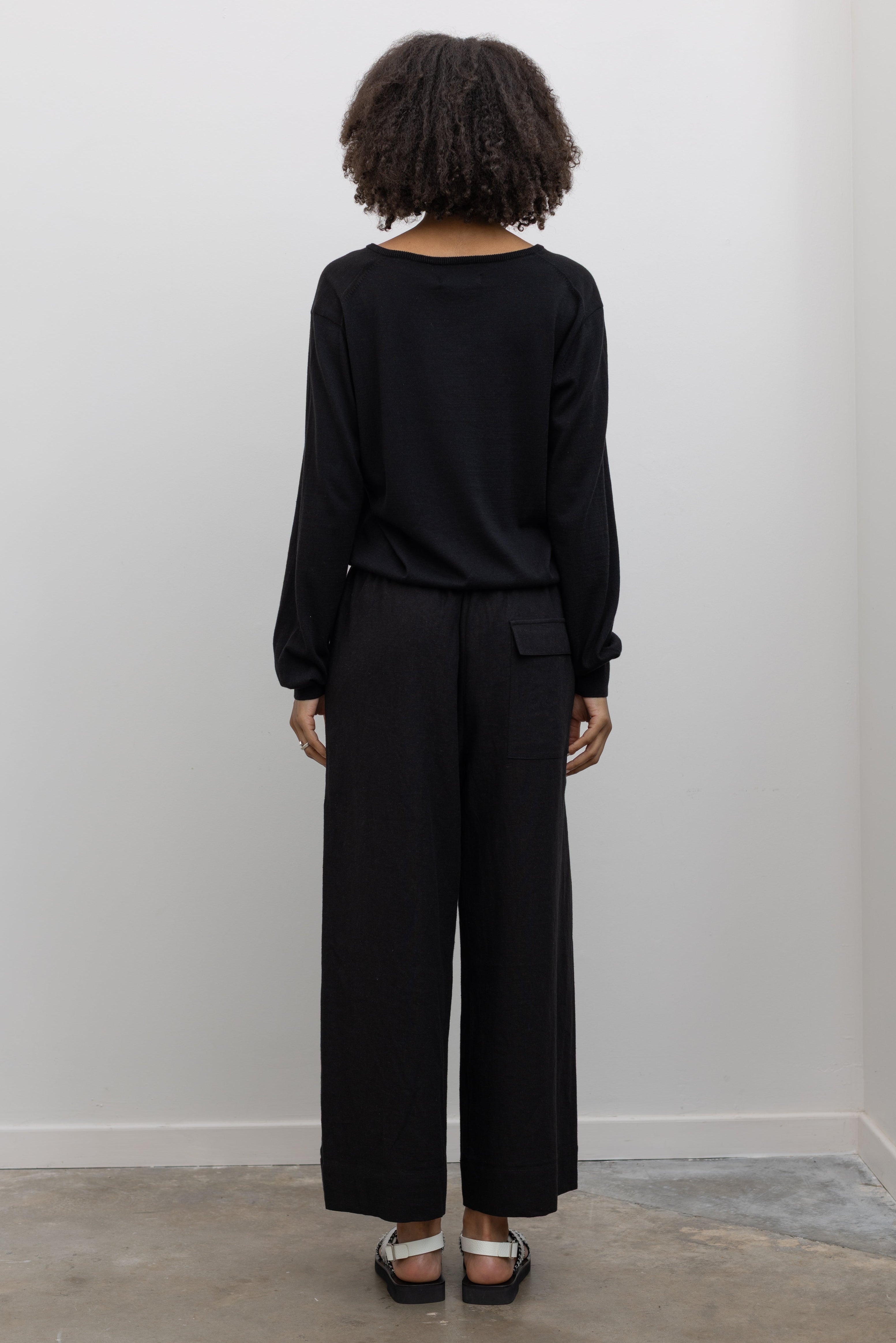 Full length back view of a woman standing wearing elevated casual weekend fashion in a Mijeong park Black Cotton Blend V-Neck Long Sleeve Knit Top paired with Mijeong Park Black Linen Blend Cropped Pants featuring a comfortable drawstring waist, front and back patch pockets, and a cropped length suitable for a minimalist, versatile wardrobe at an Alas Eius store.