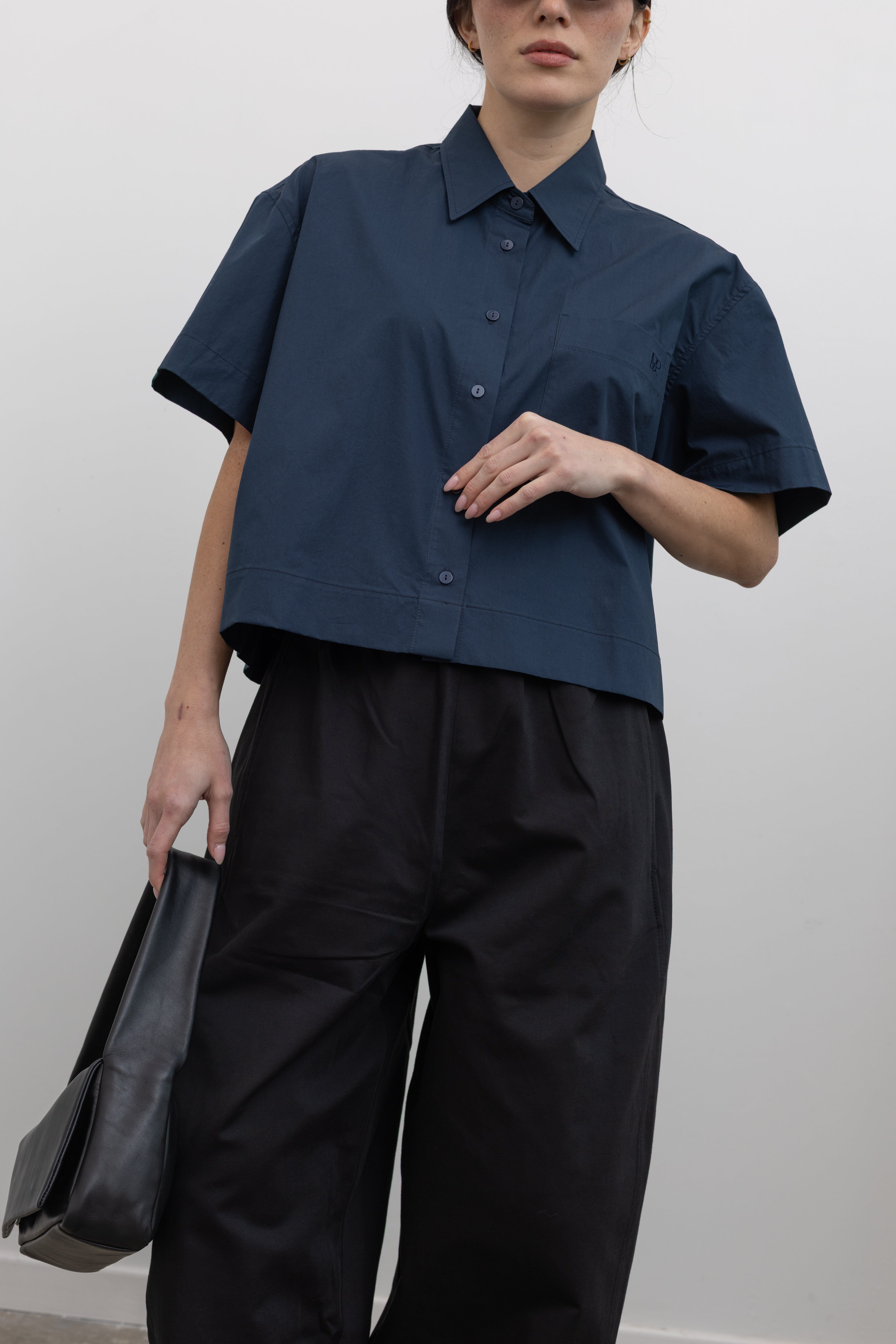 Front view of modern womenswear spring fashion in Mijeong Park black baggy balloon pants with curved outseams, a relaxed fit, elastic waistband, and back patch pockets paired with a structured navy blue short sleeve cropped hem button down shirt and a black leather St. Agni bag at an Alas Eius store. 