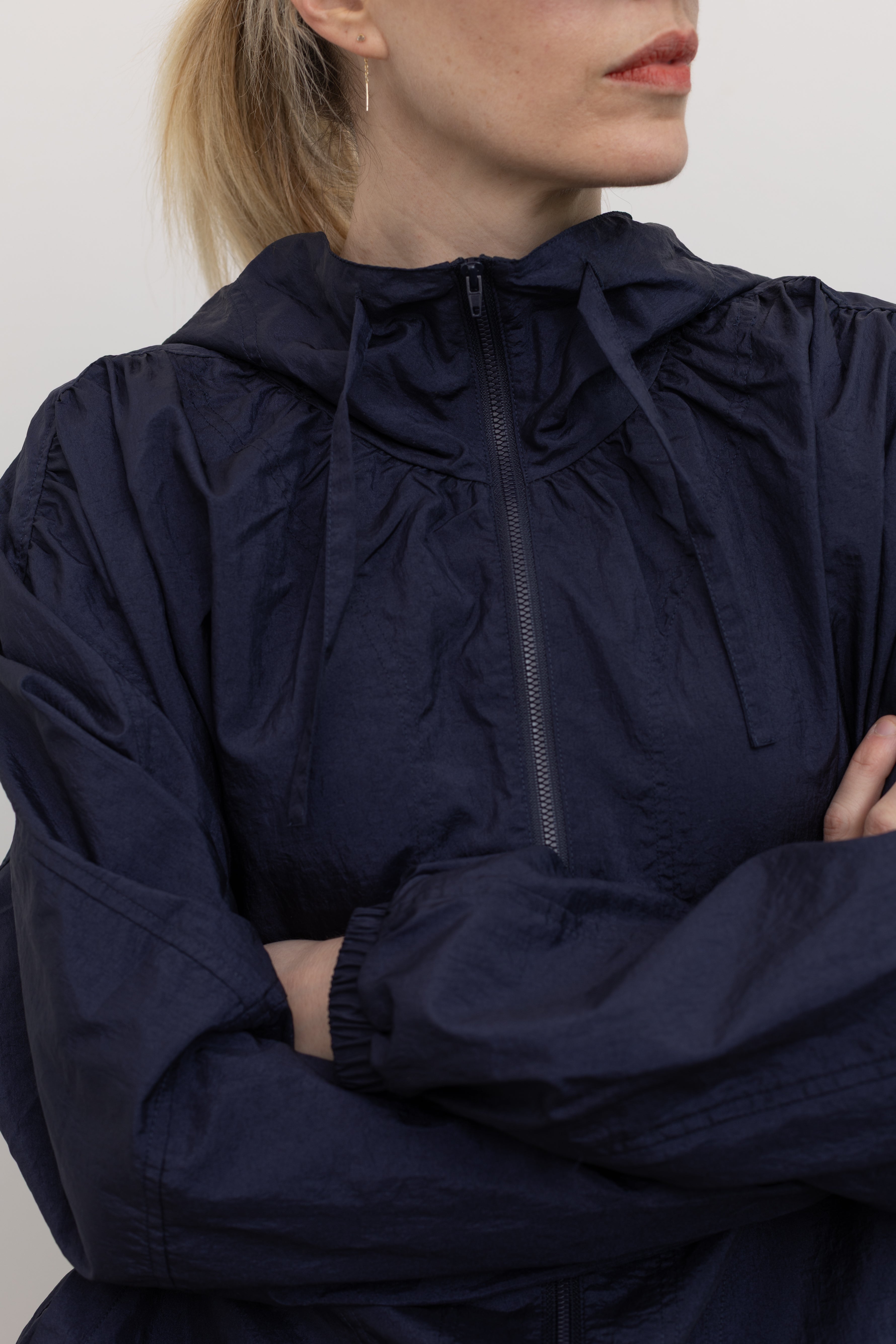 Close-up detail front view of a woman posing wearing an versatile outerwear staple in a  Mijeong Park navy blue nylon shirred anorak jacket featuring an oversized fit, front pockets, hood, and a zipper closure at an Alas Eius store. 