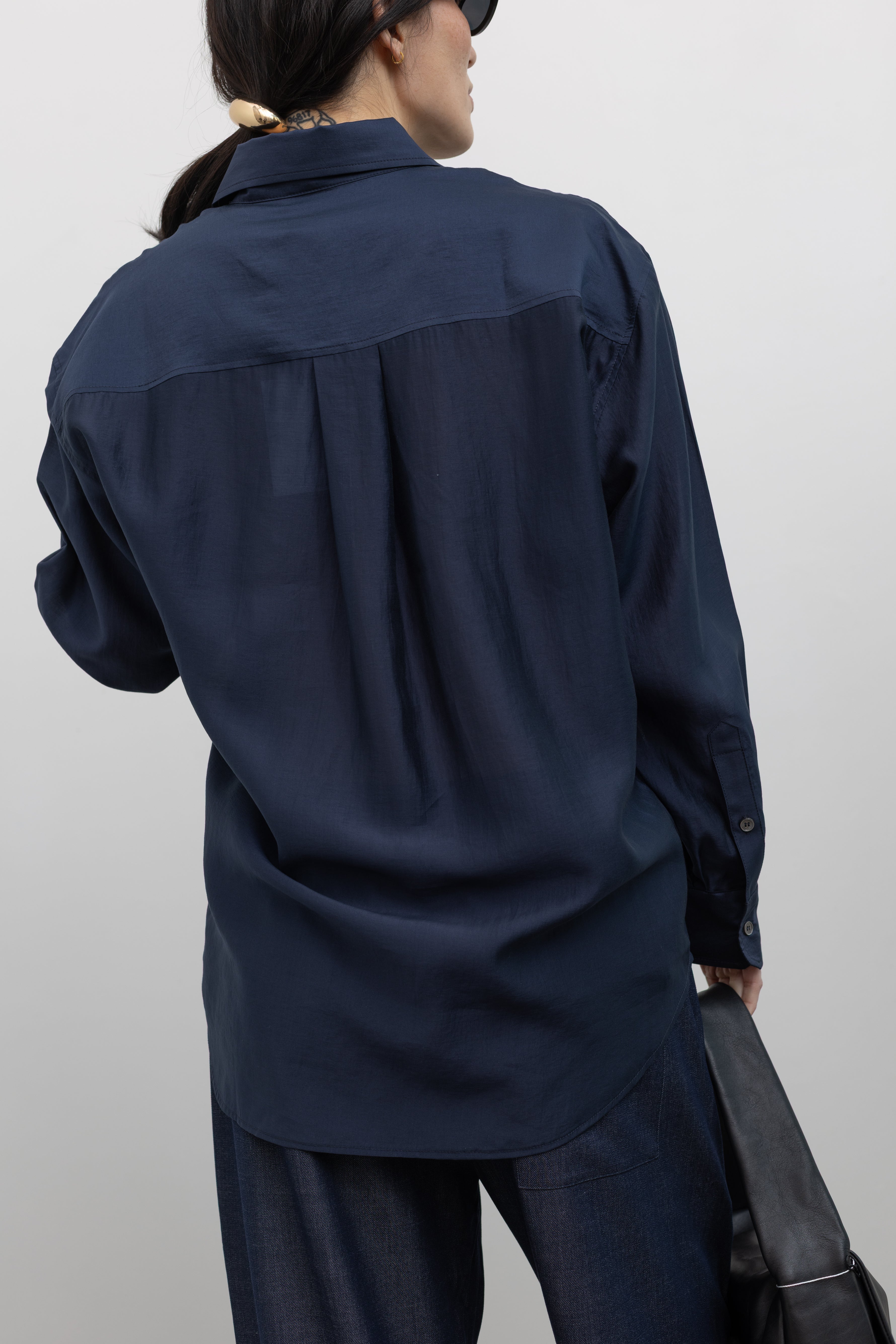 Close-up back view of a woman showing the side button detail on a Mijeong Park navy blue semi-sheer oversized shirt featuring a flattering off-center button front, long sleeves, and a classic sharp collar at an Alas Eius store. 