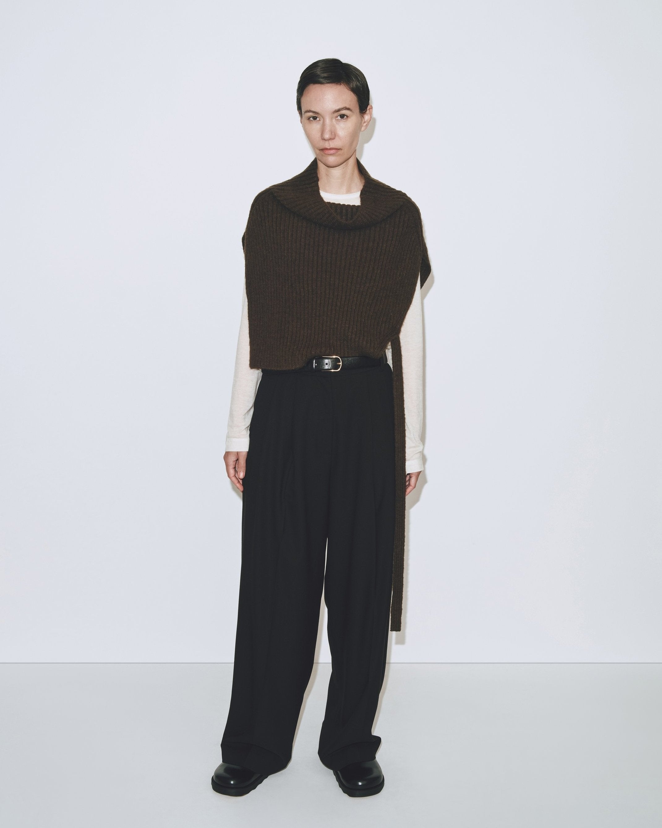 A person wearing an olive brown ribbed knitted Mijeong Park neck warmer with a mock neck neckline and tie waist detail, paired with a black trousers at Alas Eius