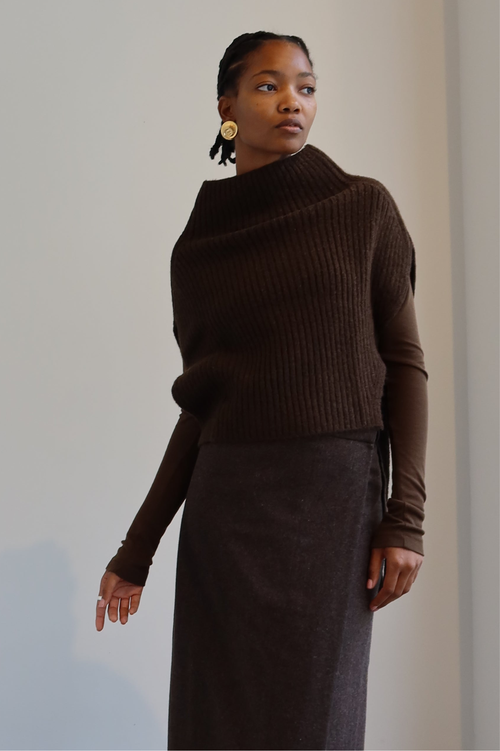 A person wearing an olive brown ribbed knitted Mijeong Park neck warmer with a mock neck neckline and tie waist detail, paired with a brown skirt at Alas Eius
