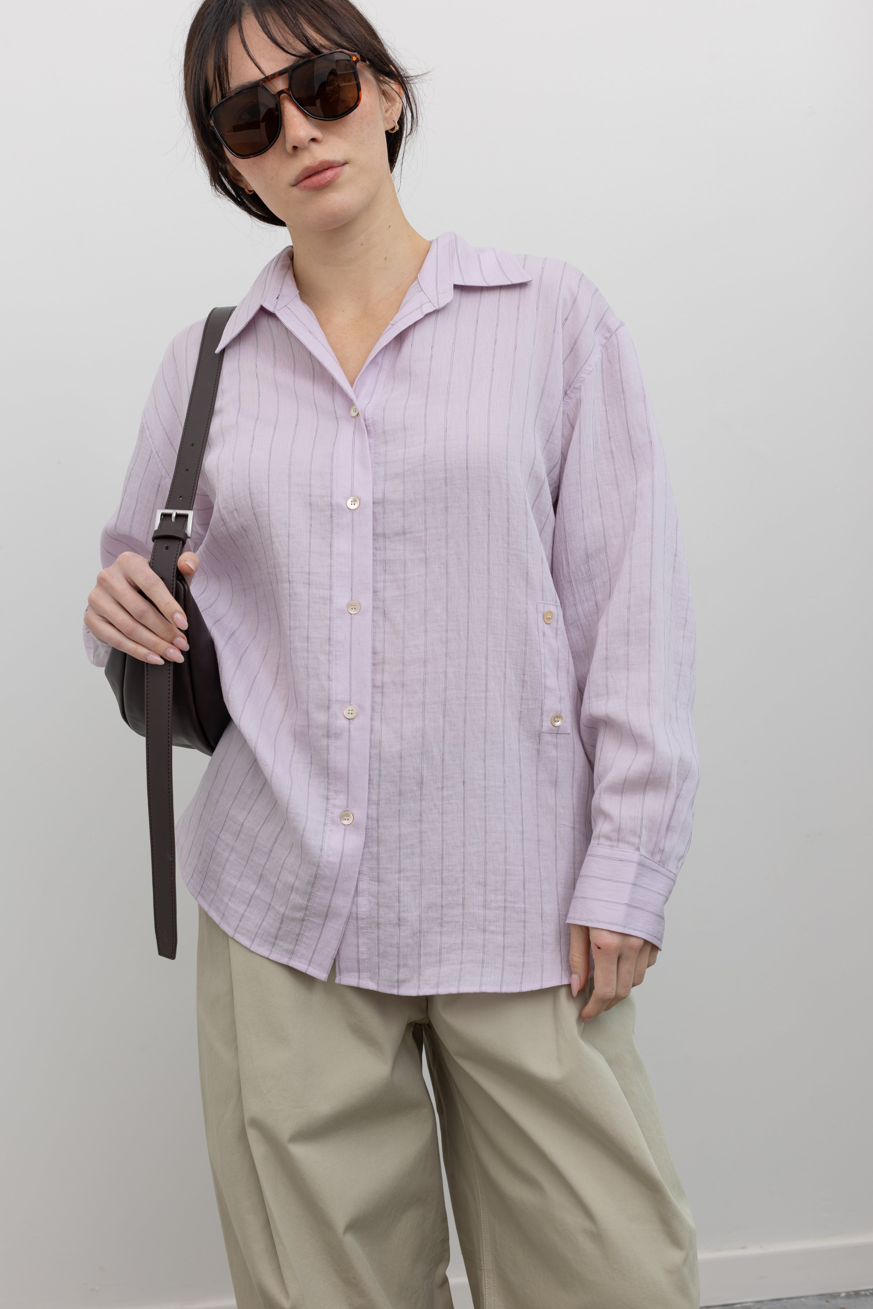 Close-up front view of a woman wearing elevated weekend and travel capsule wardrobe staples in a Mijeong Park oversized soft pink pinstripe button down shirt featuring a classic collar, long sleeves, and an off center button down front paired with an elevated classic khaki baggy curved leg pant with strappy sandals at an Alas Eius store. 