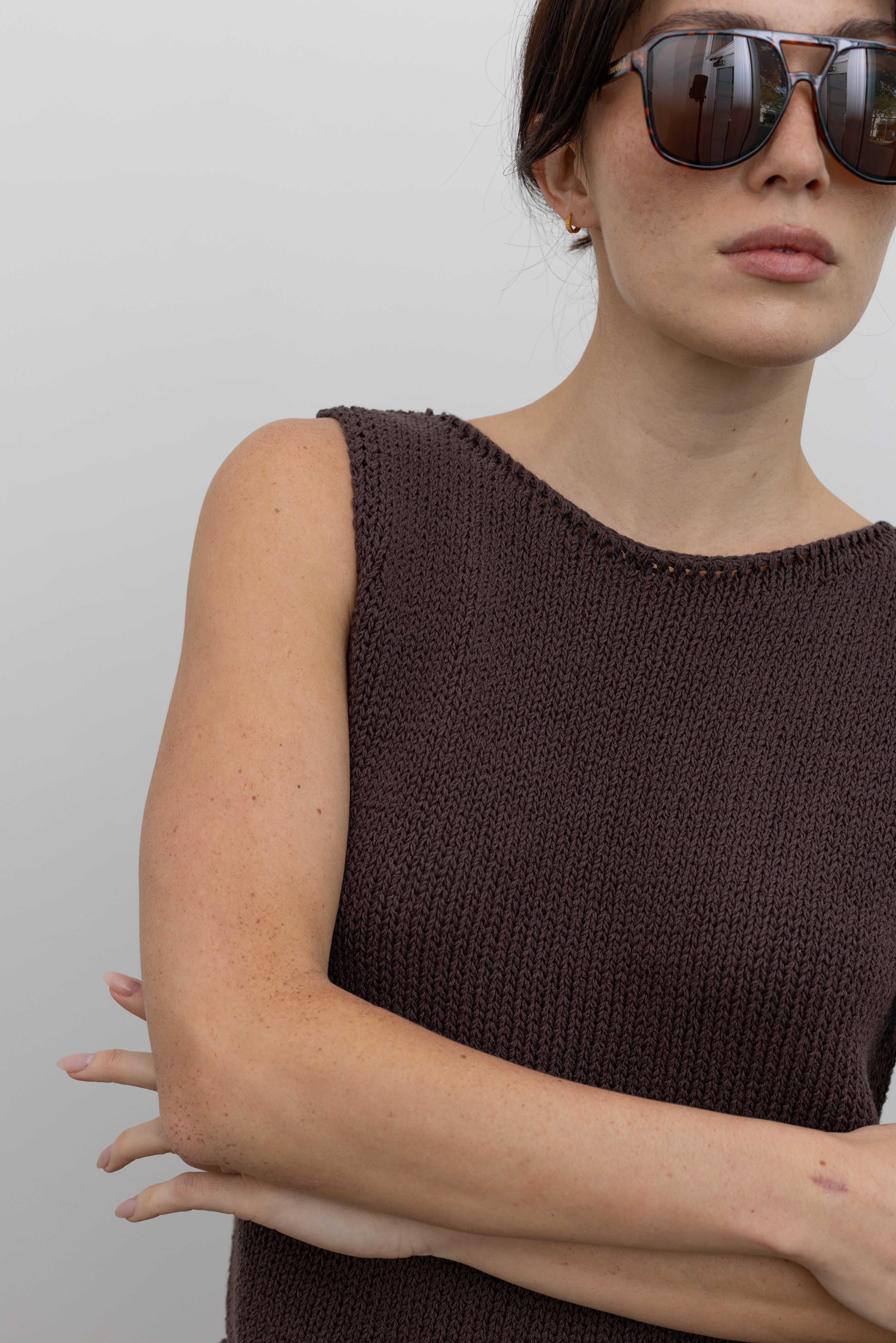 Close-up front view of a woman posing wearing modern minimalist spring and summer fashion in a Mijeong Park dark brown boat neck knit tank featuring a relaxed fit and rolled edges at the neck and hem at an Alas Eius store.  