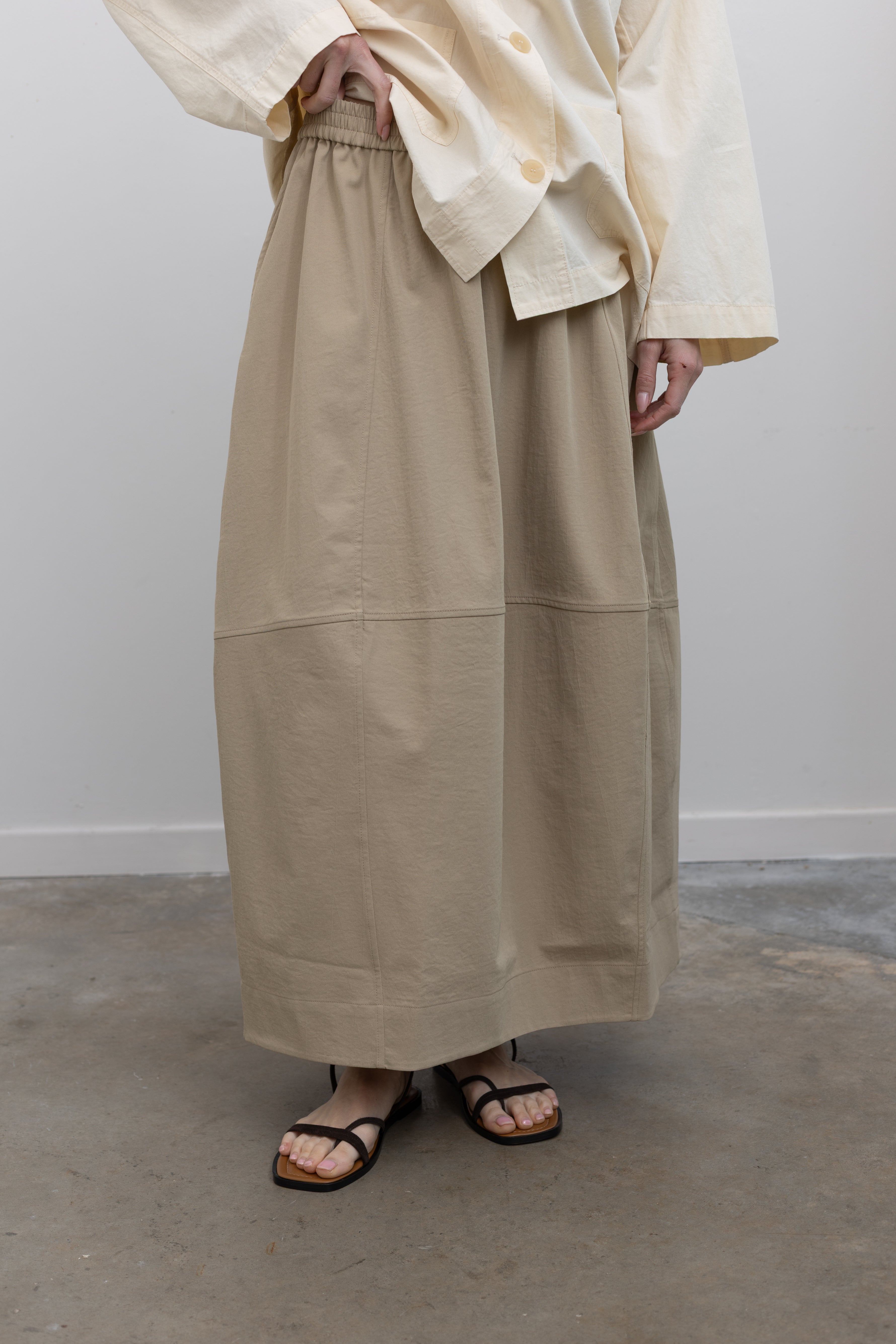 Full length front view of a woman standing wearing contemporary womenswear spring fashion in a Mijeong Park cream colored button down work jacket with a beige voluminous balloon midi-to-maxi silhouette skirt featuring an elastic waistband paired with black thin strappy sandals at an Alas Eius store. 