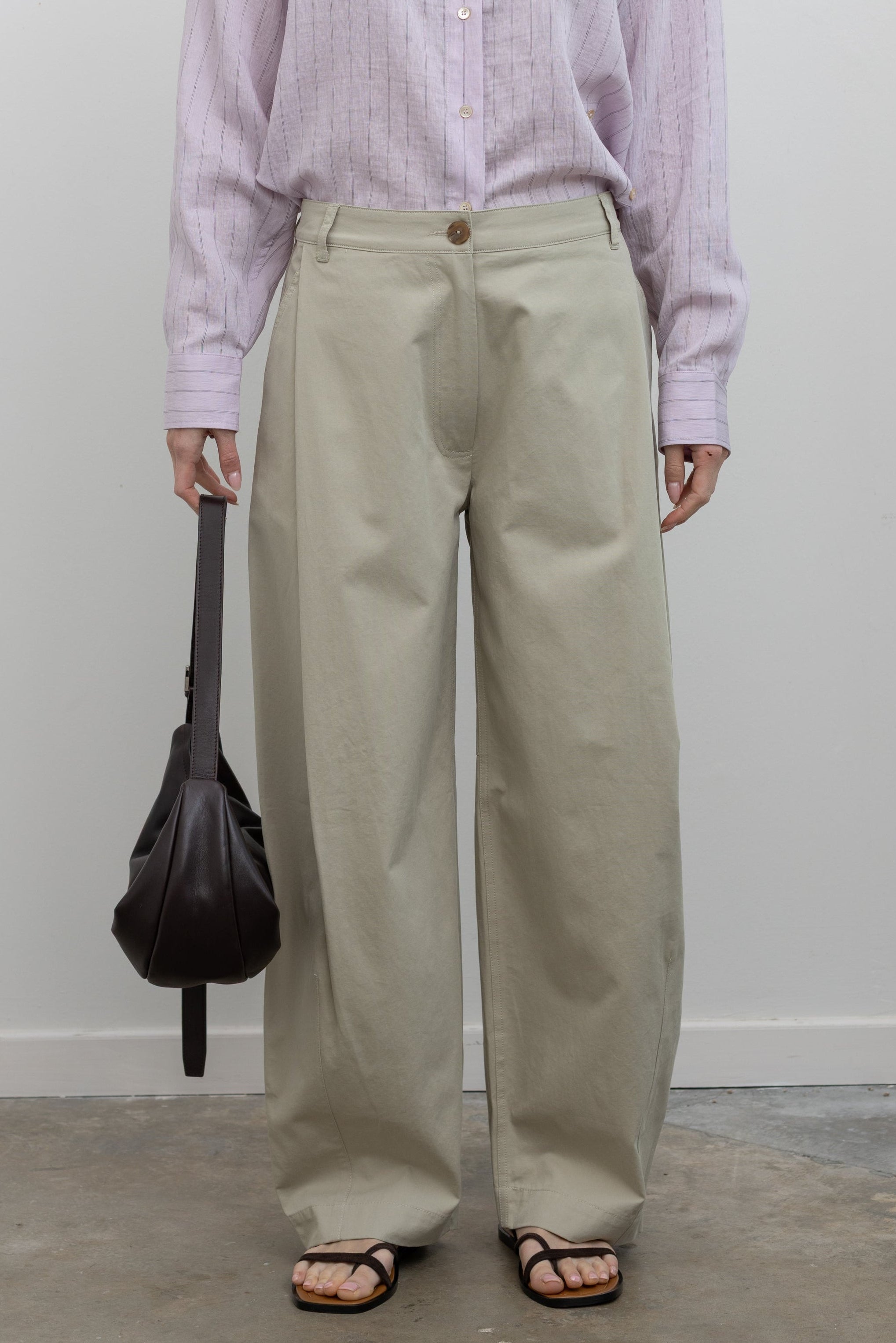 Full length front view of a woman standing wearing sophisticated spring fashion in Mijeong Park beige 100% cotton baggy barrel leg pants featuring curved outseams that create architectural volume and a partially elasticated waistband and belt loops for a tailored look paired with an elevated classic Mijeong Park pink stripe button down shirt with a St. Agni leather bag and strappy sandals at an Alas Eius store. 