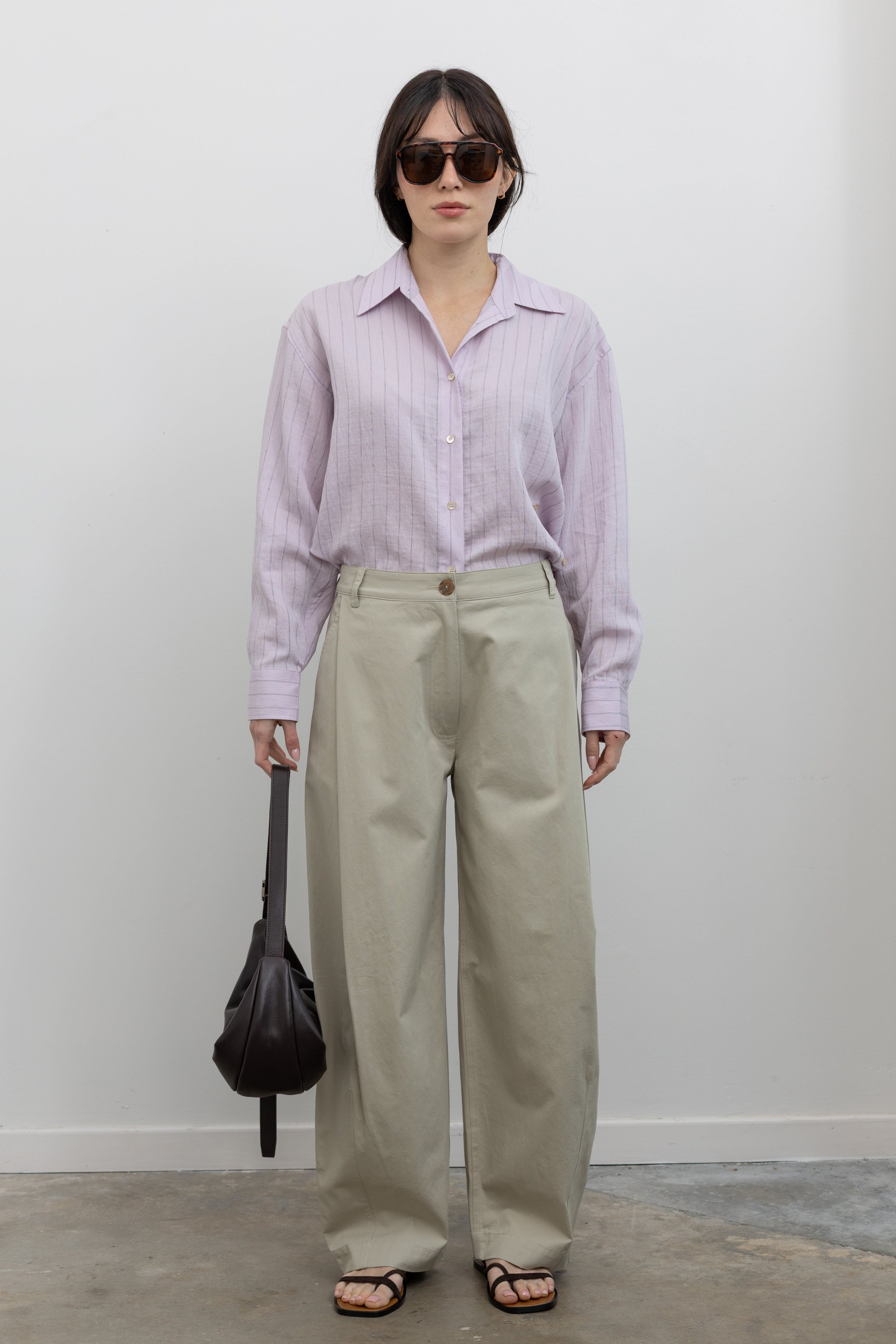 Full length front view of a woman standing wearing sophisticated spring fashion in Mijeong Park beige 100% cotton baggy barrel leg pants featuring curved outseams that create architectural volume and a partially elasticated waistband and belt loops for a tailored look paired with an elevated classic Mijeong Park pink stripe button down shirt with a St. Agni leather bag and strappy sandals at an Alas Eius store. 