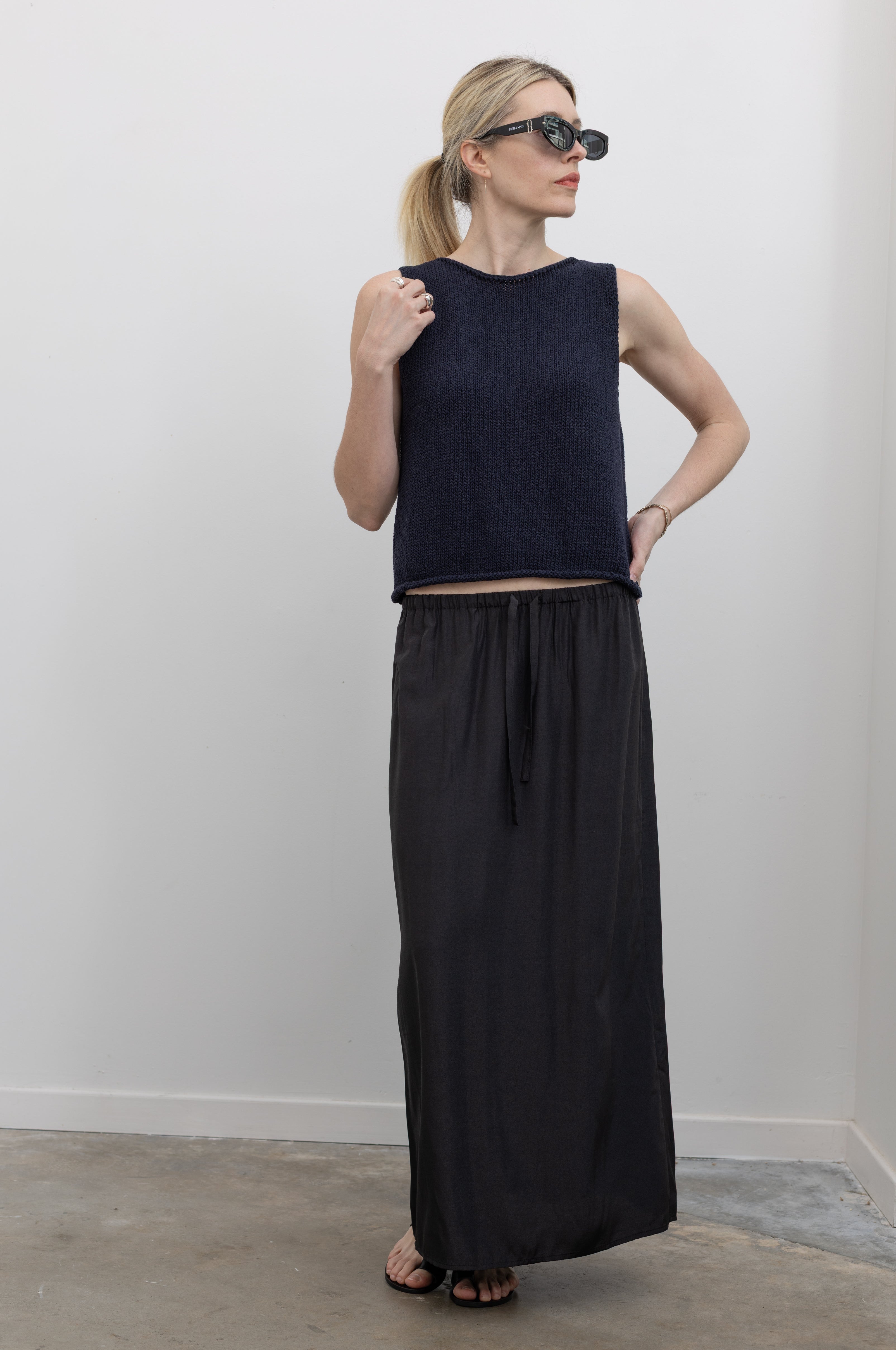 Full length front view of a woman wearing chic and modern spring fashion in a Mijeong Park black midi to maxi length skirt featuring an elastic drawstring waistband and discreet back slit paired with a Mijeong Park navy blue boat neck knit tank and strappy sandals at an Alas Eius store. 