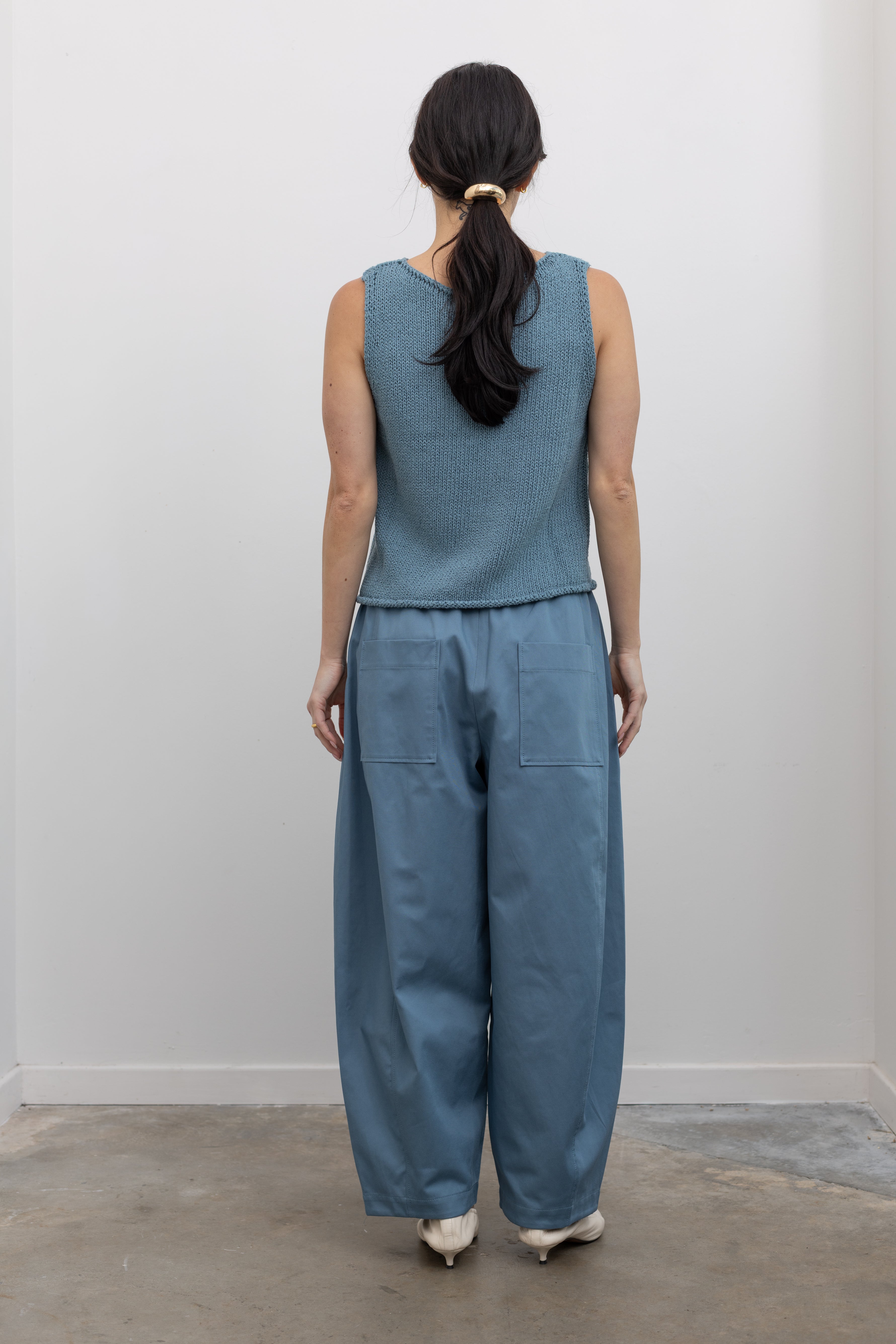 Full length back view of a woman wearing spring fashion in a Mijeong Park light blue boat neck knit sweater tank top featuring rolled edges around the neckline and hem at an Alas Eius store. 