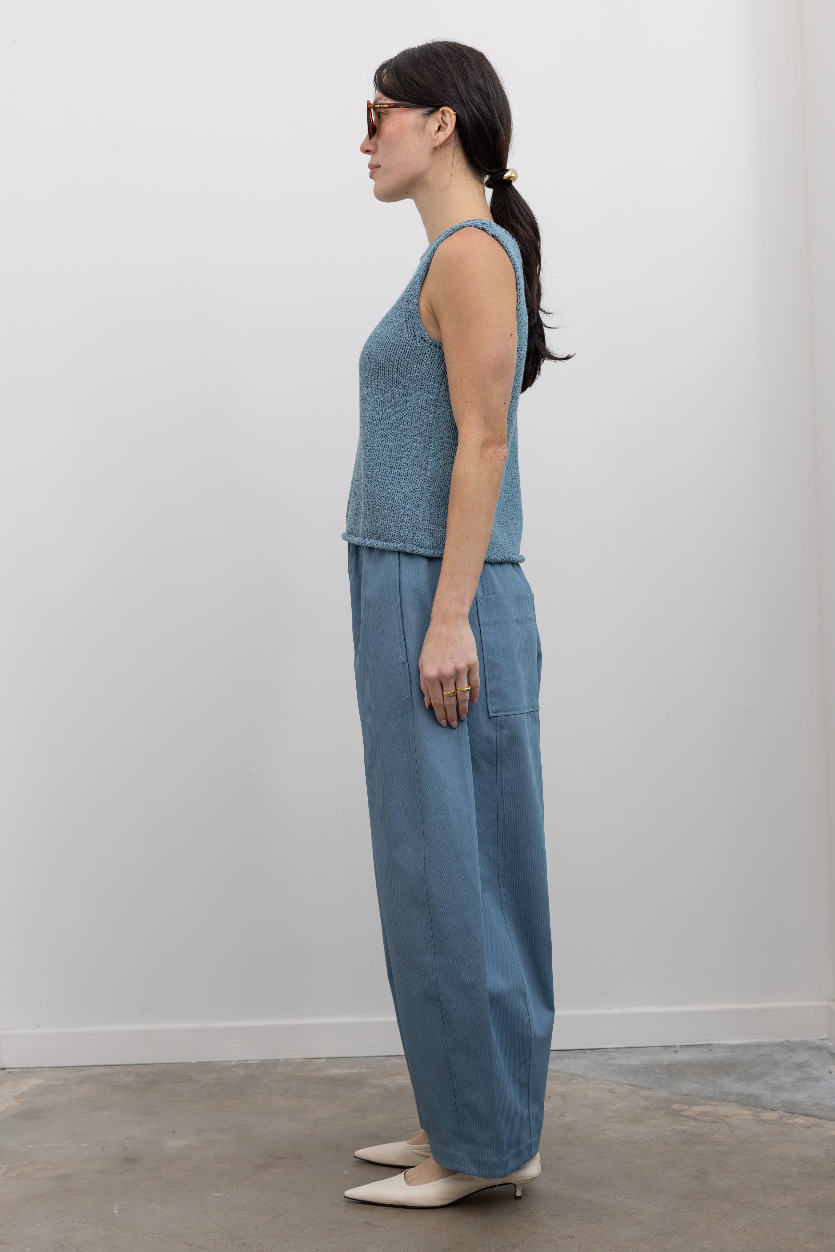 Full length side view of a woman wearing spring monochromatic fashion in Mijeong Park light blue cropped balloon pants with curved outseams, a relaxed fit, mid-rise waist, and a comfortable elasticated waistband paired with a Mijeong Park blue boat neck knit tank with white leather kitten heels at an Alas Eius store. 