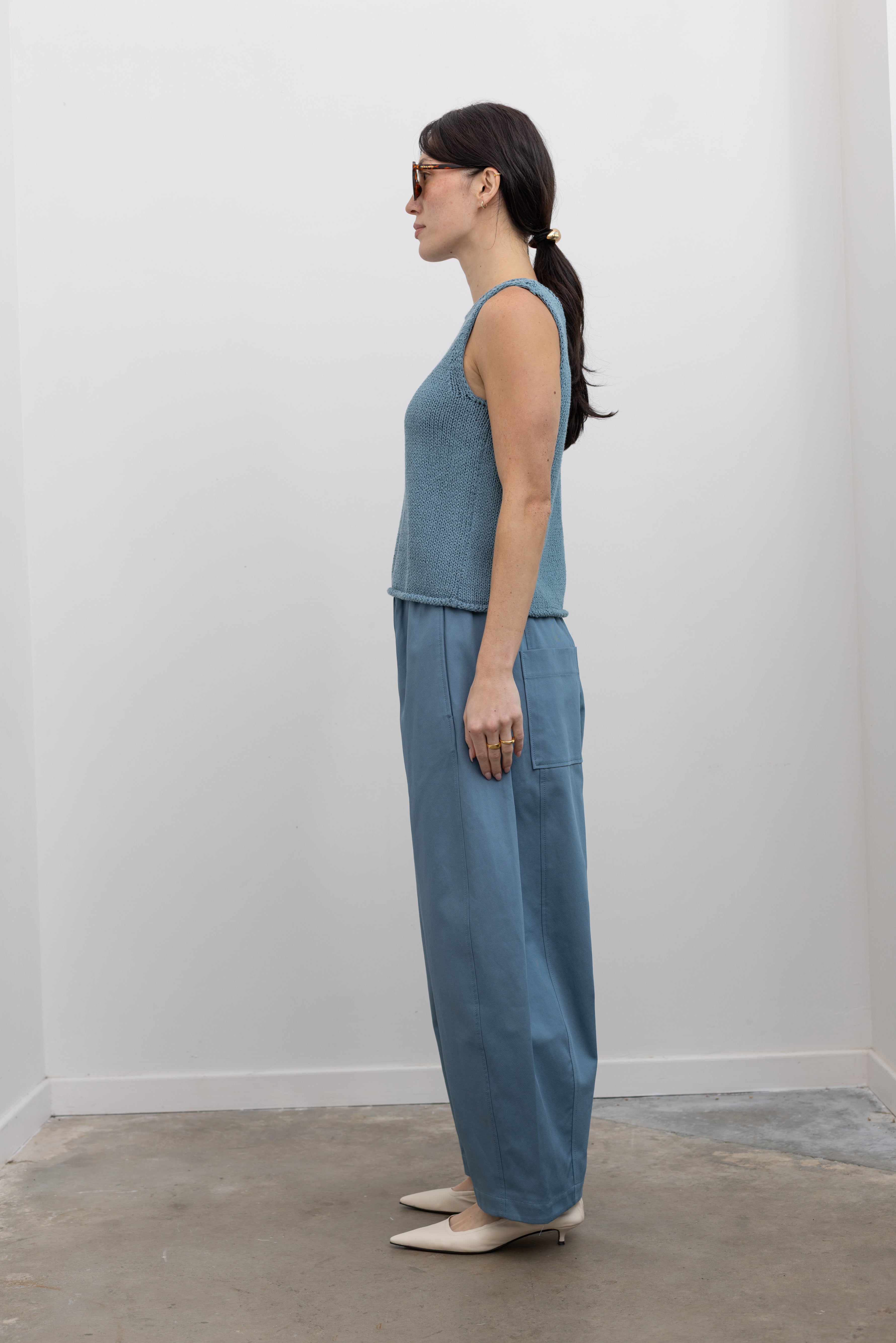 Full length side view of a woman wearing chic and modern spring fashion in a Mijeong Park light blue boat neck knit sweater tank top featuring rolled edges around the neckline and hem at an Alas Eius store. 