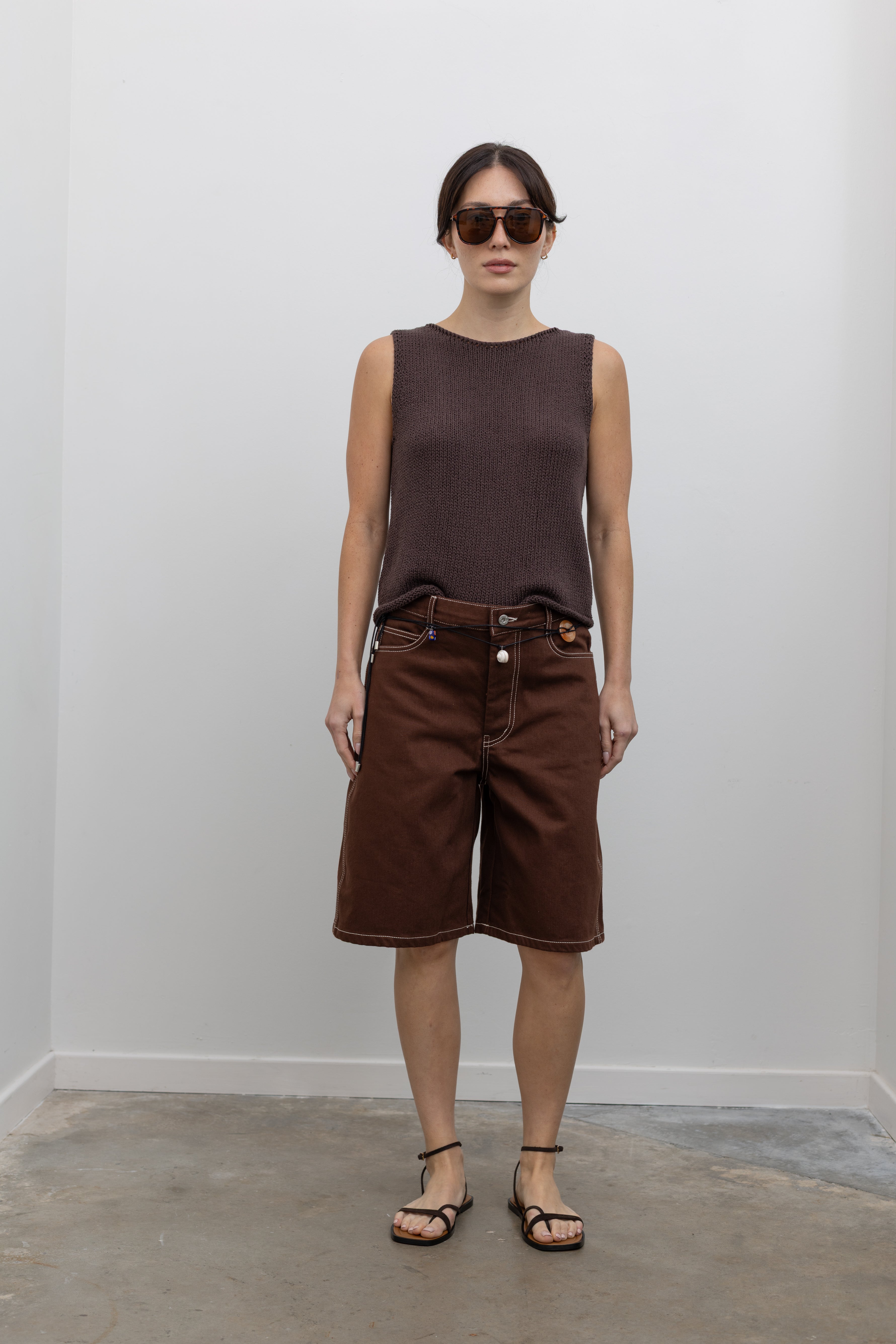 Full length front view of a woman posing wearing modern minimalist spring and summer fashion in a Mijeong Park dark brown boat neck knit tank featuring a relaxed fit and rolled edges at the neck and hem paired with Kowtow Framework Organic Cotton Brown Denim Long Shorts that sit just above the knee and feature white contrast stitching at an Alas Eius store.  