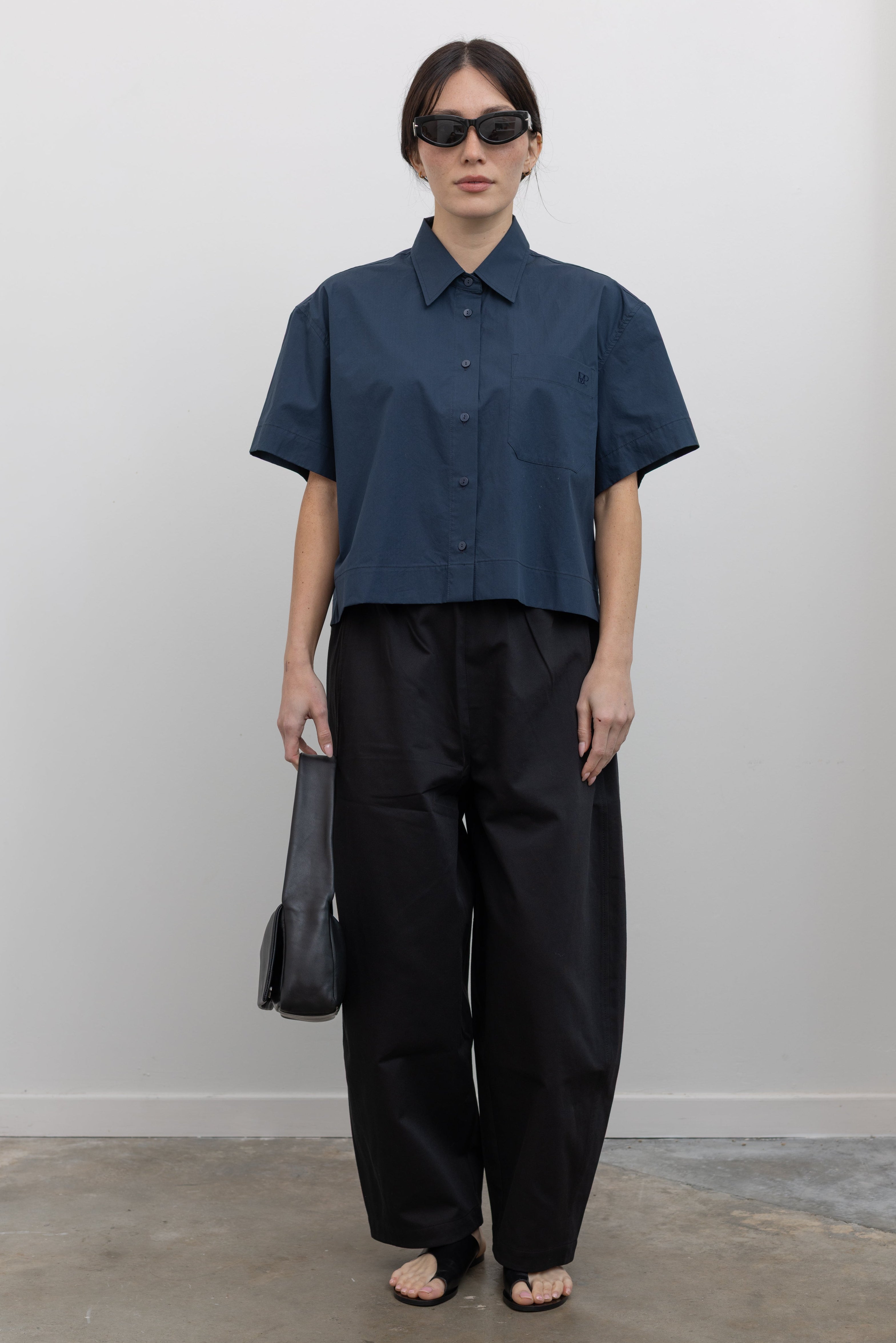 Front view of contemporary womenswear spring fashion in Mijeong Park black baggy balloon pants with curved outseams, a relaxed fit, elastic waistband, and back patch pockets paired with a structured navy blue short sleeve cropped hem button down shirt and a black leather St. Agni bag at an Alas Eius store. 