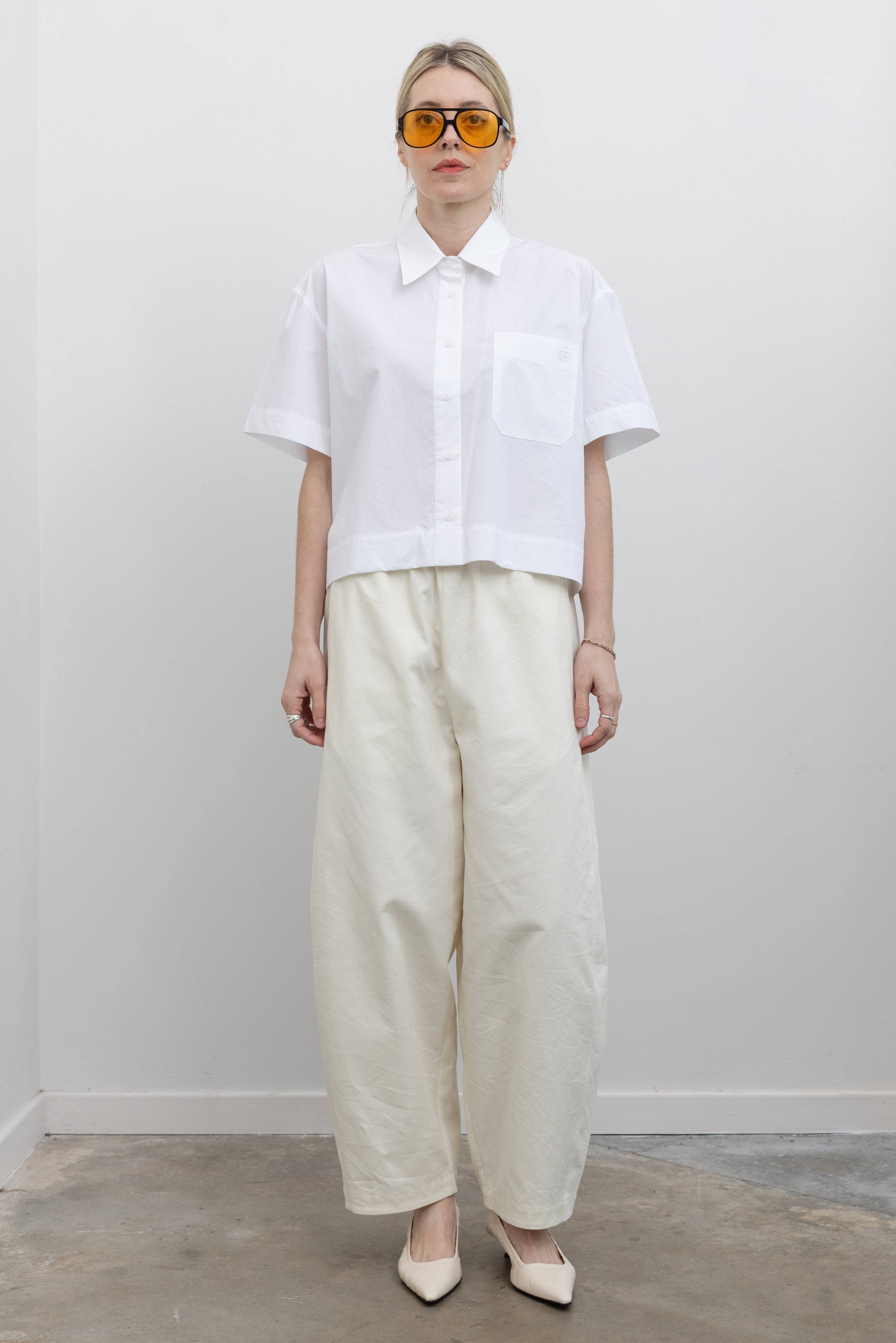 Full length front view of a woman wearing spring monochromatic fashion in Mijeong Park cream colored cropped balloon pants with curved outseams, 
a relaxed fit, mid-rise waist, and a comfortable elasticated band paired with a Mijeong Park crisp white cotton oversized cropped collared shirt with a front pocket at an Alas Eius store. 