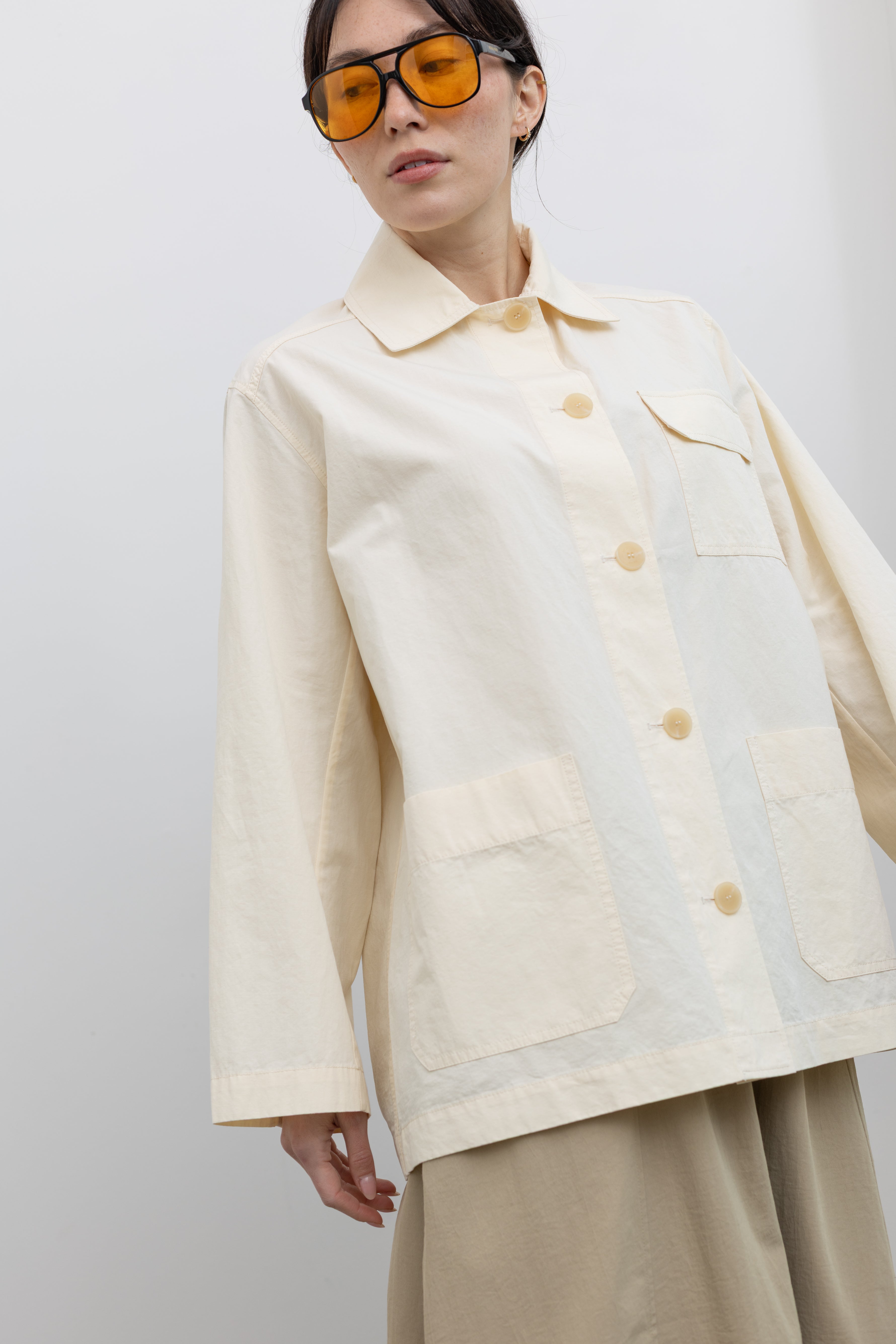 Close-up front view of a woman posing wearing chic and modest Spring fashion in a Mijeong Park beige balloon midi skirt paired with a cream colored linen blend work jacket featuring a relaxed fit, two front patch pockets and a button down front closure for versatile styling at an Alas Eius store. 