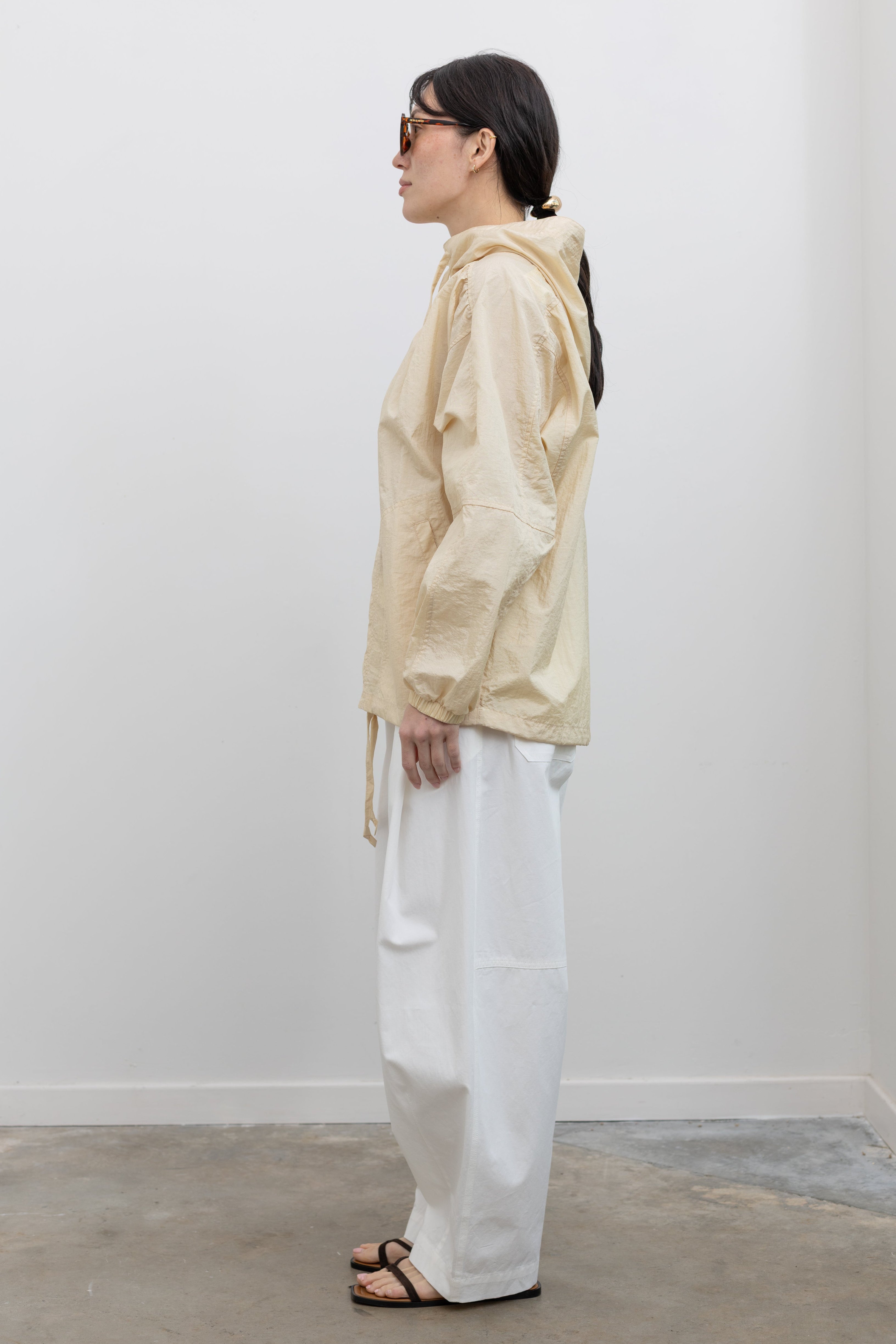 Full length side view of a woman wearing an elevated minimalist outerwear staple in a Mijeong Park cream colored nylon shirred anorak jacket featuring an oversized fit, front pockets, hood, and a zipper closure paired with white curved leg pants at an Alas Eius store. 