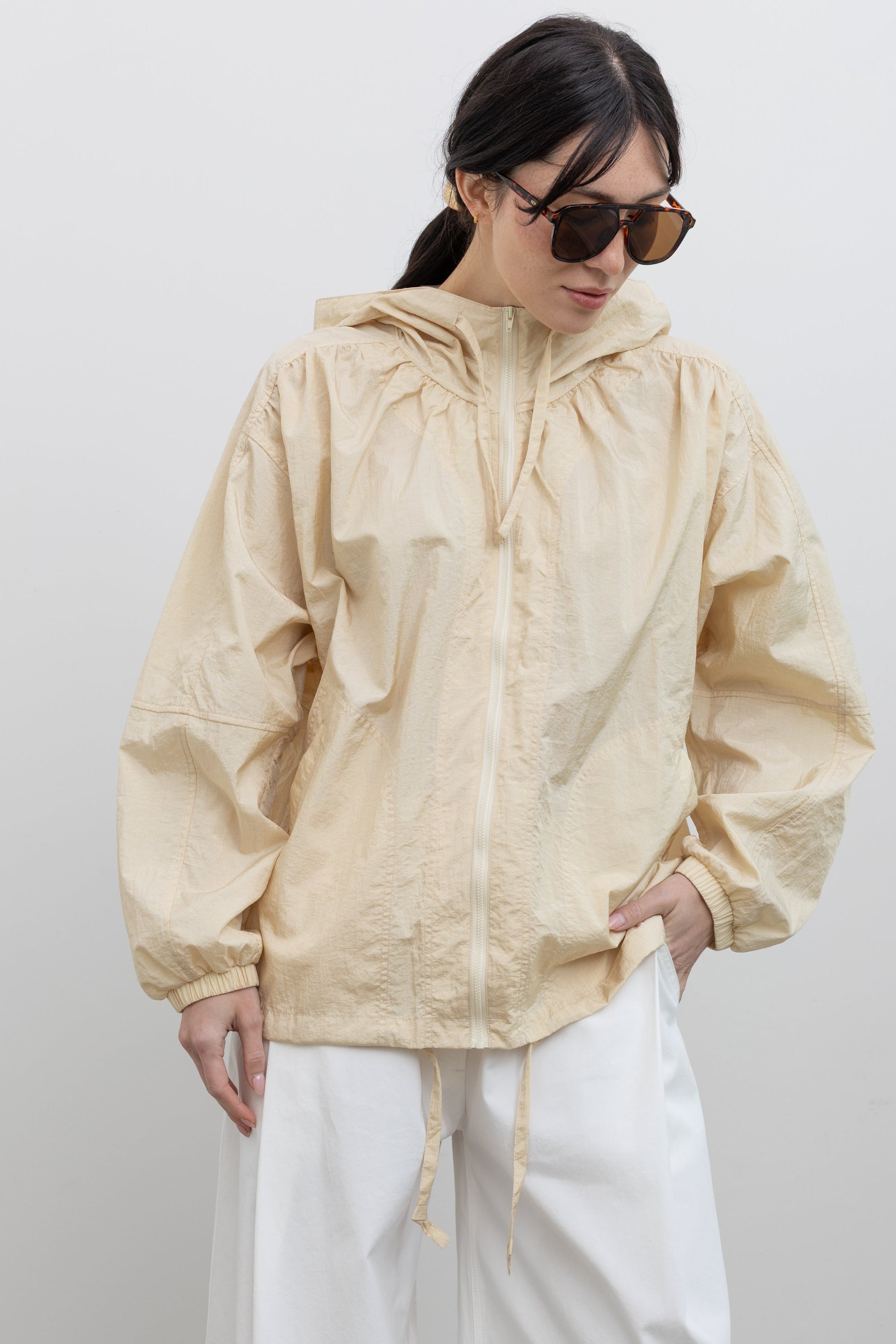 Close-up front view of a woman posing with sunglasses wearing a versatile outerwear staple in a Mijeong Park cream colored nylon shirred anorak jacket featuring an oversized fit, front pockets, hood, and a zipper closure at an Alas Eius store. 