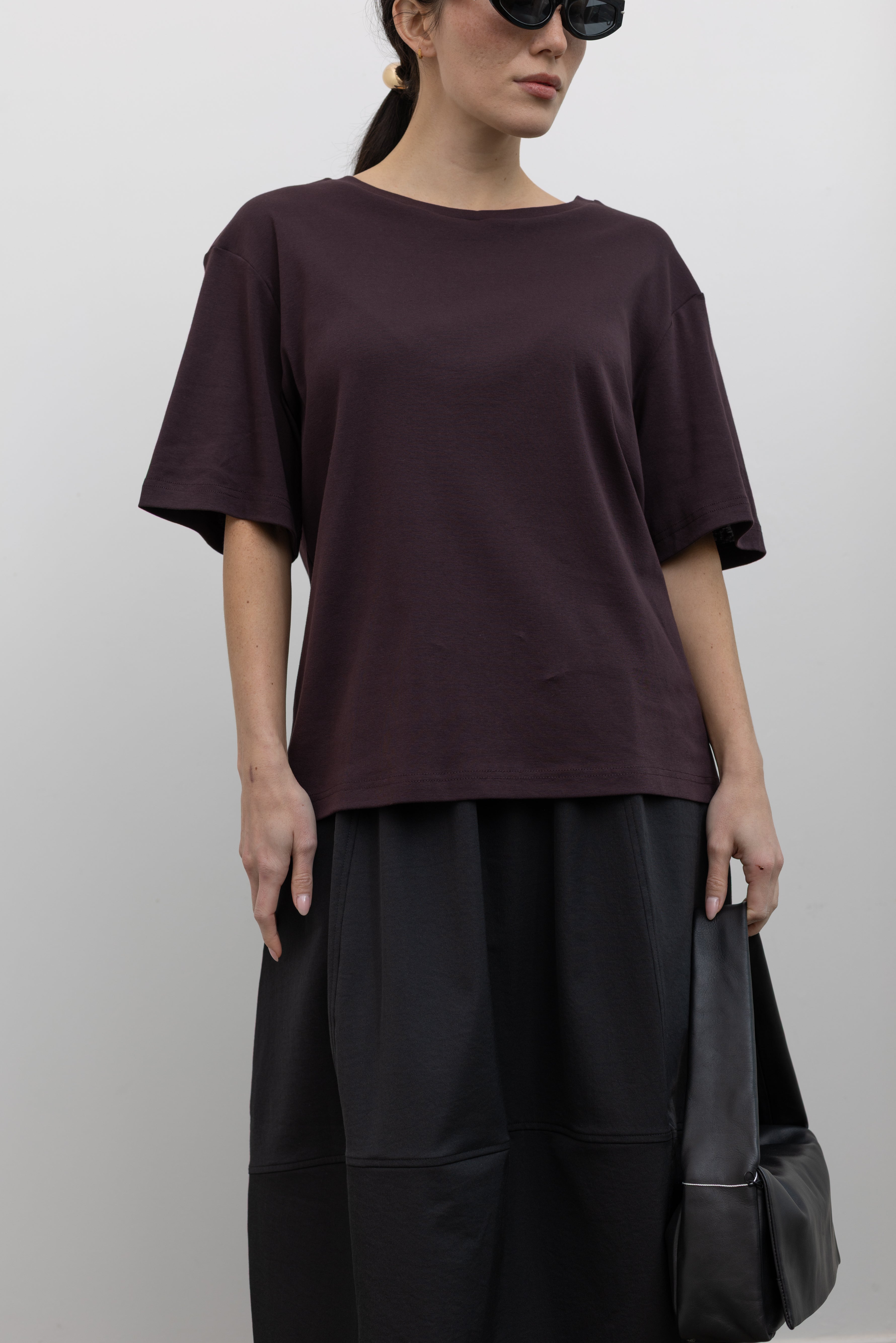 Front view of a woman standing wearing elevated essential spring fashion in a Mijeong Park dark brown oversized t-shirt with a dropped shoulder paired with an Alas Eius label black nylon skirt and a modern leather St. Agni bag at an Alas Eius store. 