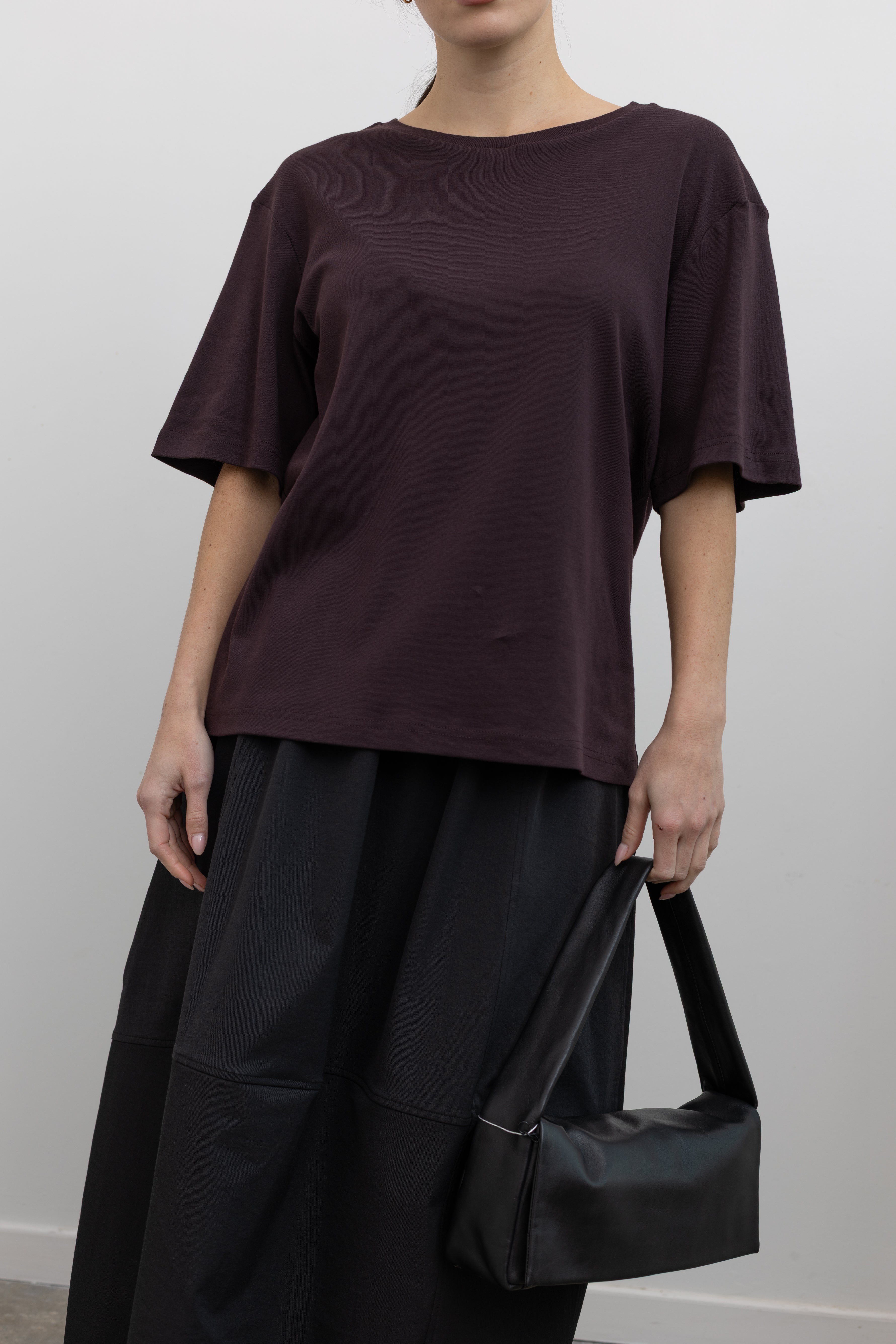 Front view of a woman standing wearing elevated minimalist spring fashion in a Mijeong Park dark brown oversized t-shirt with a dropped shoulder paired with an Alas Eius label black nylon skirt and a modern leather St. Agni bag at an Alas Eius store. 