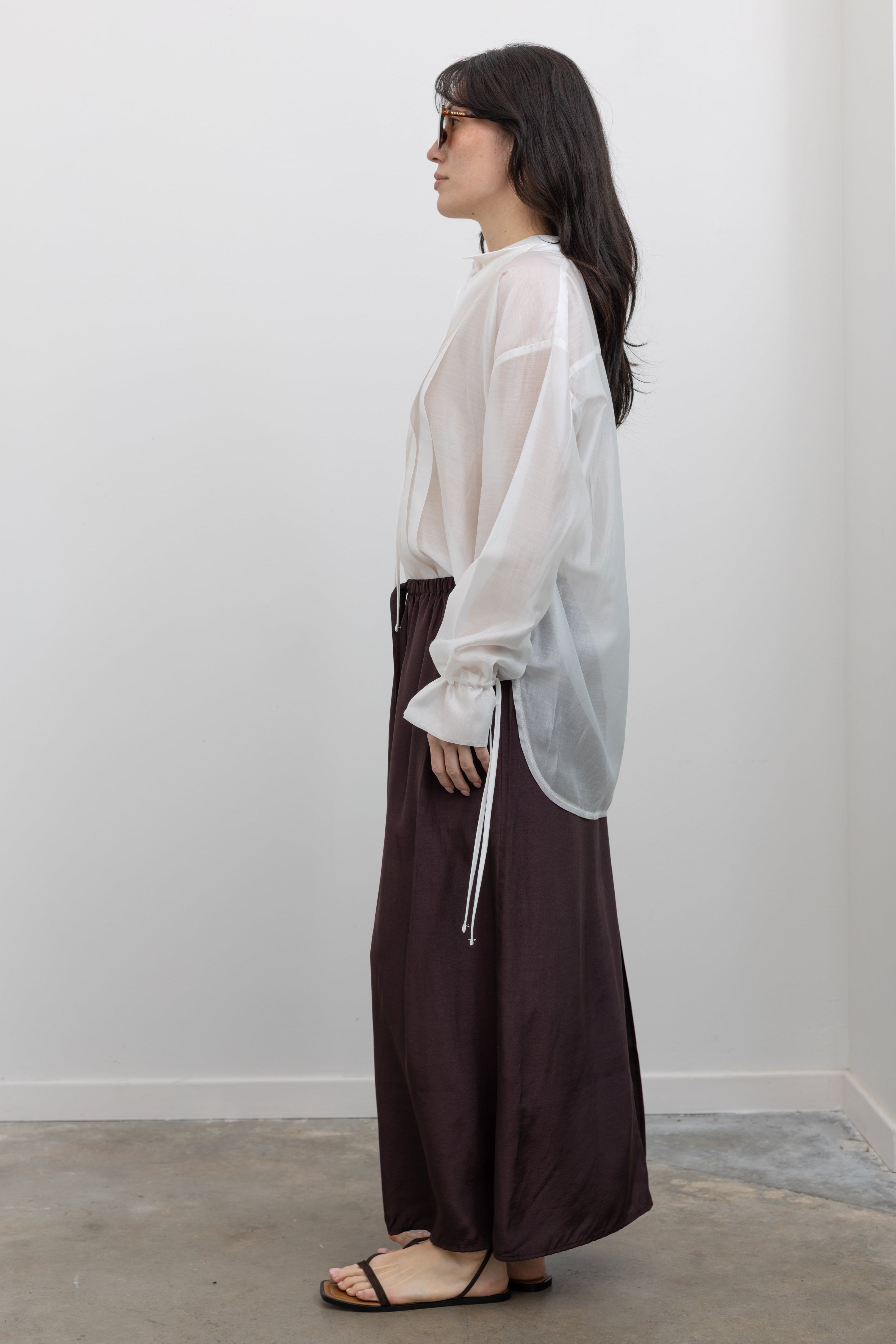 Full length side view of a woman wearing spring fashion in a chic and versatile Mijeong Park dark brown midi to maxi length skirt featuring an elastic drawstring waistband and discreet back slit paired with Collective brand white organza sheer blouse and black strappy sandals at an Alas Eius store. 
