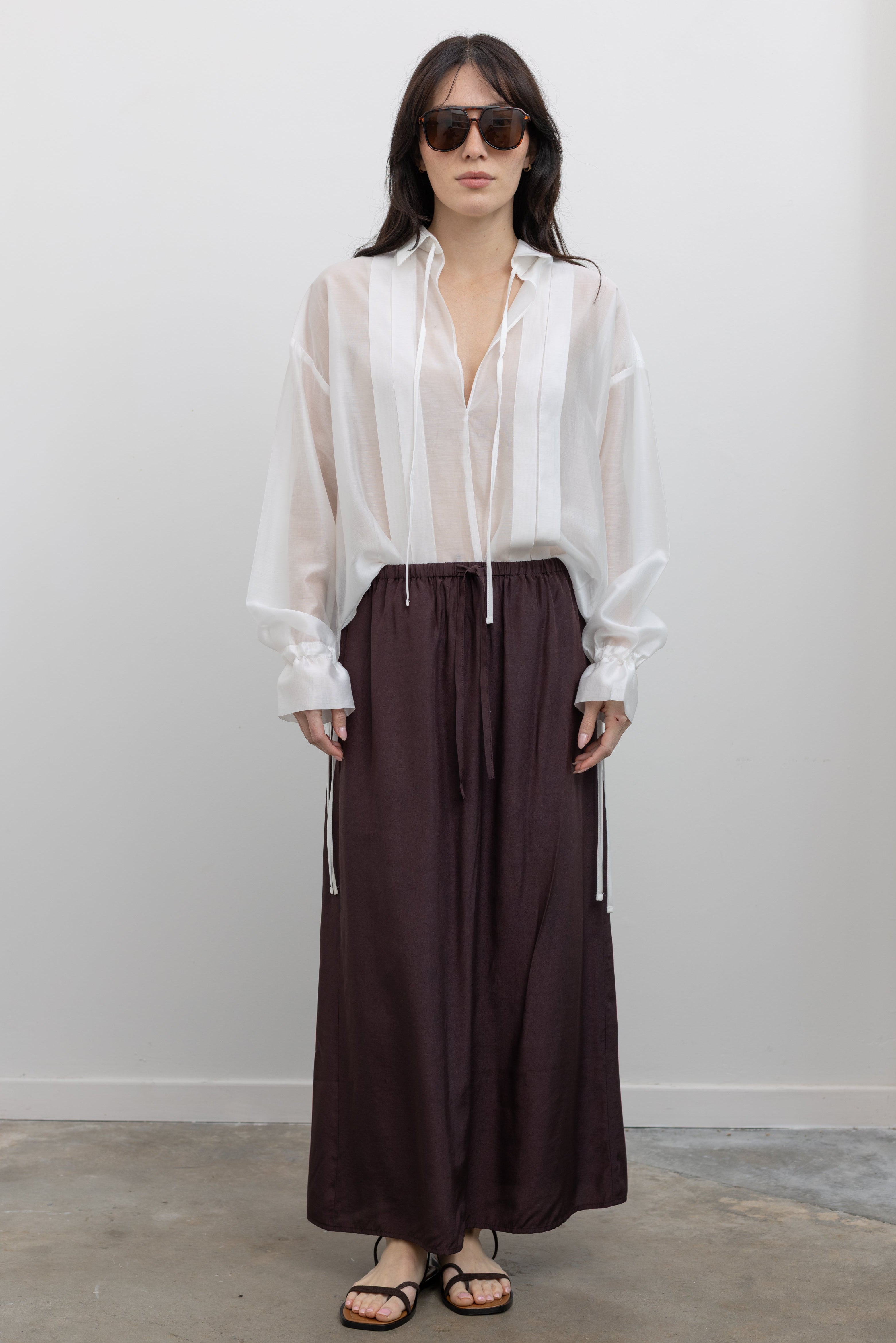 Full length front view of a woman wearing spring fashion in a chic and versatile Mijeong Park dark brown midi to maxi length skirt featuring an elastic drawstring waistband and discreet back slit paired with Collective brand white organza sheer blouse and black strappy sandals at an Alas Eius store. 