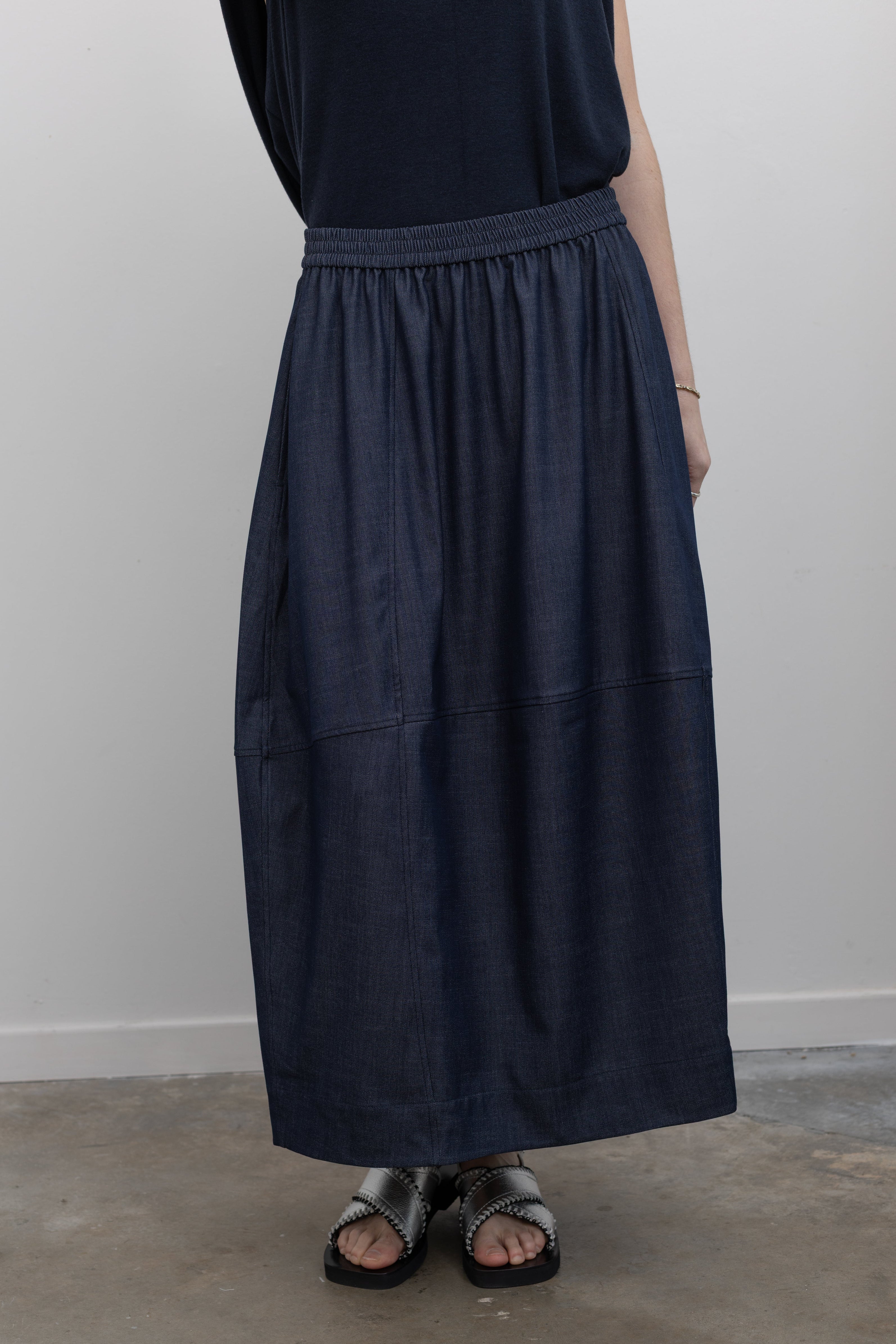 Close-up front view of a woman standing wearing a versatile yet chic Mijeong Park dark wash denim blue voluminous balloon midi-to-maxi silhouette skirt featuring an elastic waistband at Alas Eius.