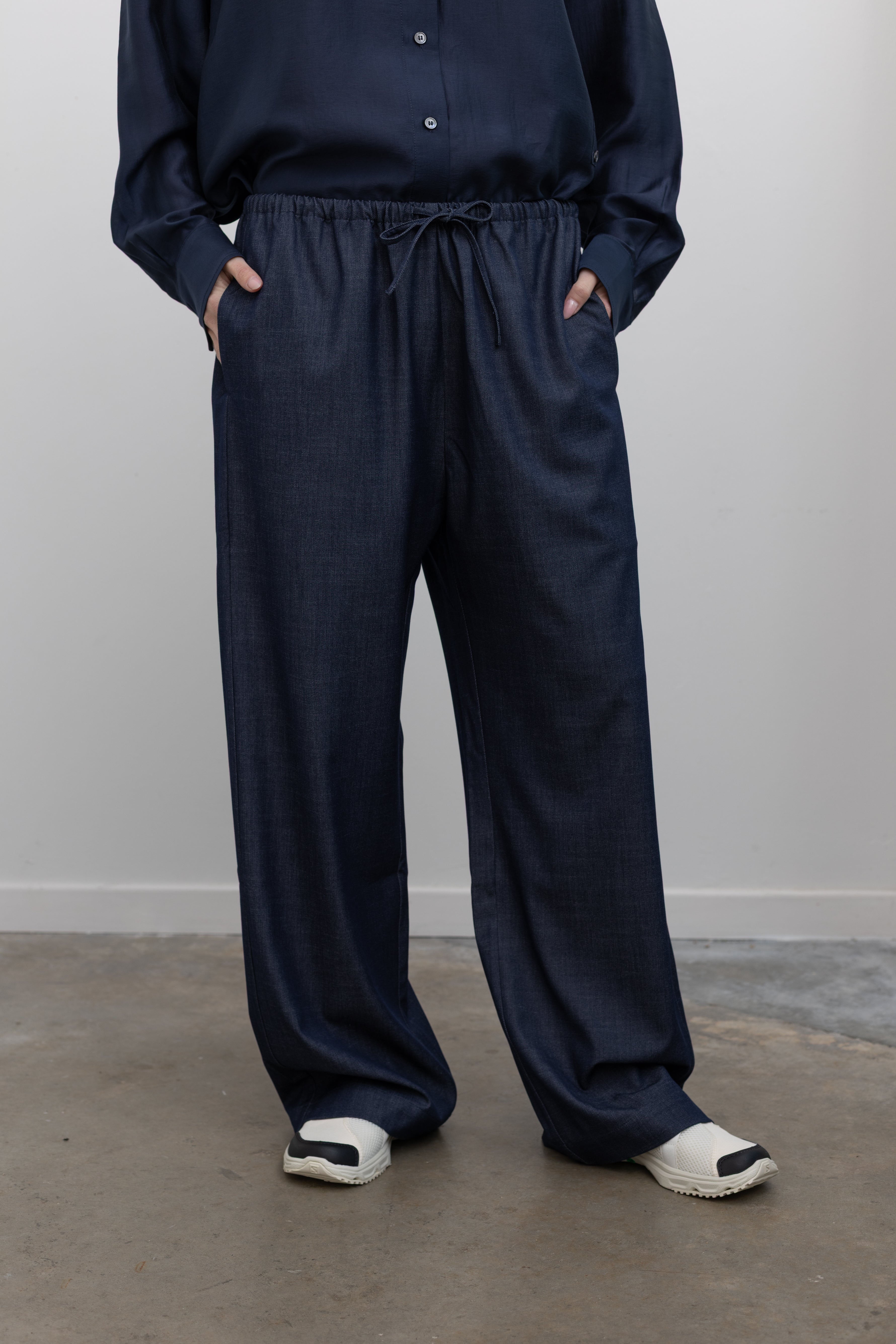 Close-up front view of a woman standing with her hands in the pockets of Mijeong Park lightweight dark wash blue denim drawstring pants featuring an elastic waistband and side seam pockets at an Alas Eius store. 

