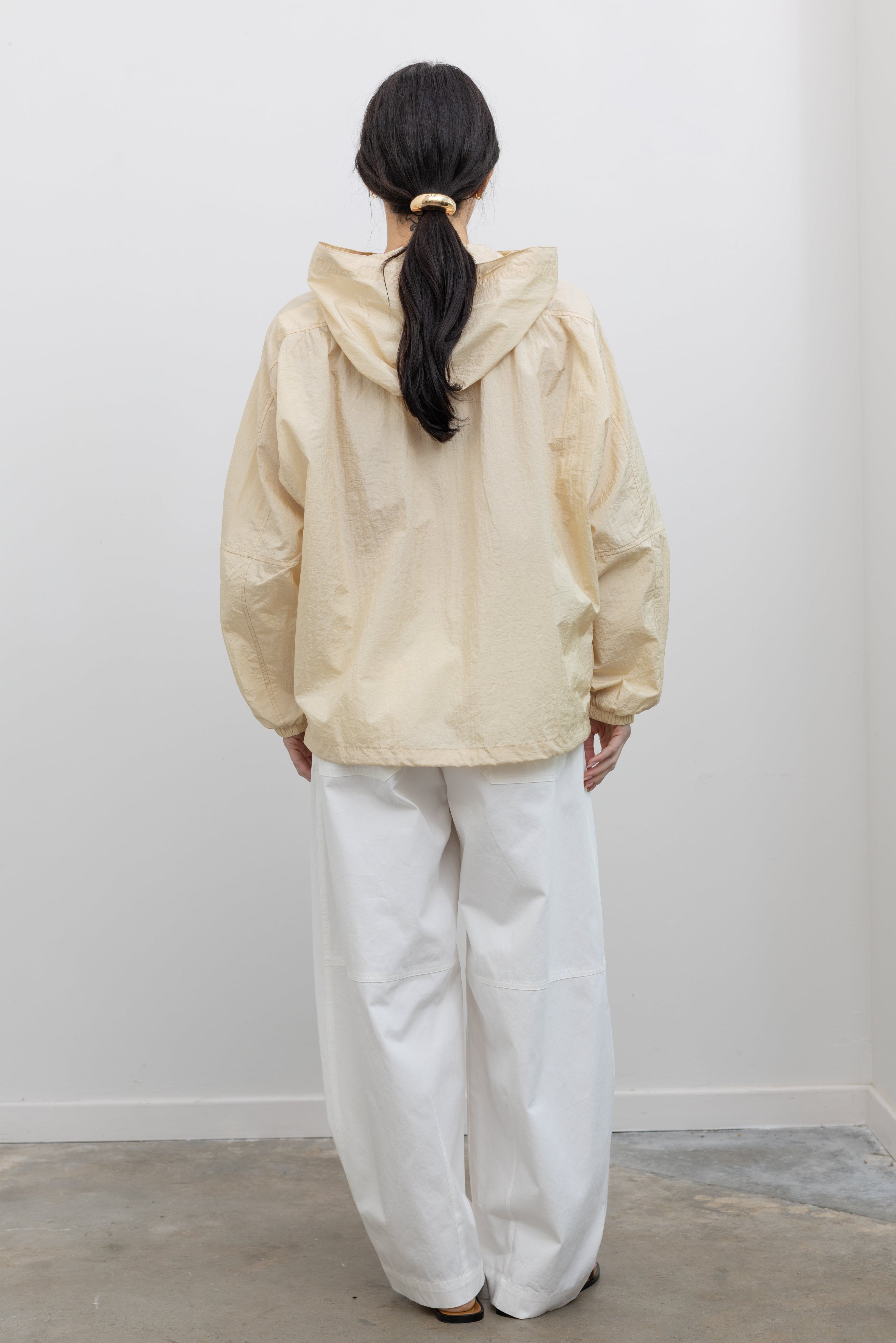 Full length back view of a woman wearing an elevated 90's inspired outerwear staple in a Mijeong Park cream colored nylon shirred anorak jacket featuring an oversized fit, front pockets, hood, and a zipper closure paired with white curved leg pants at an Alas Eius store. 