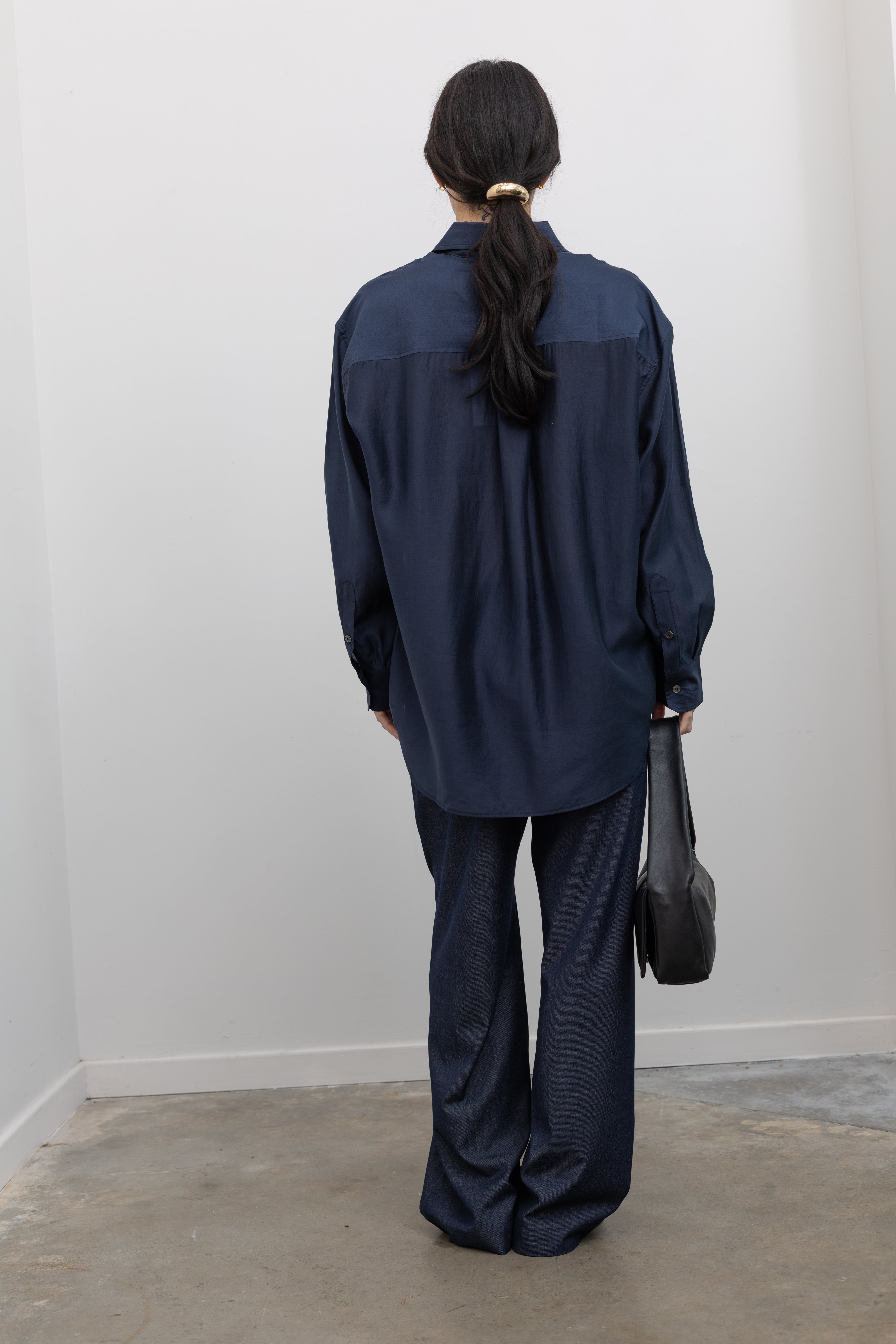 Full length back view of a woman wearing a 90's inspired staple in a Mijeong Park navy blue semi-sheer oversized shirt featuring a flattering off-center button front, long sleeves, and a classic sharp collar paired with Mijeong Park lightweight dark wash denim drawstring pants, Salomon slide sneakers, and a leather St. Agni bag at an Alas Eius store. 