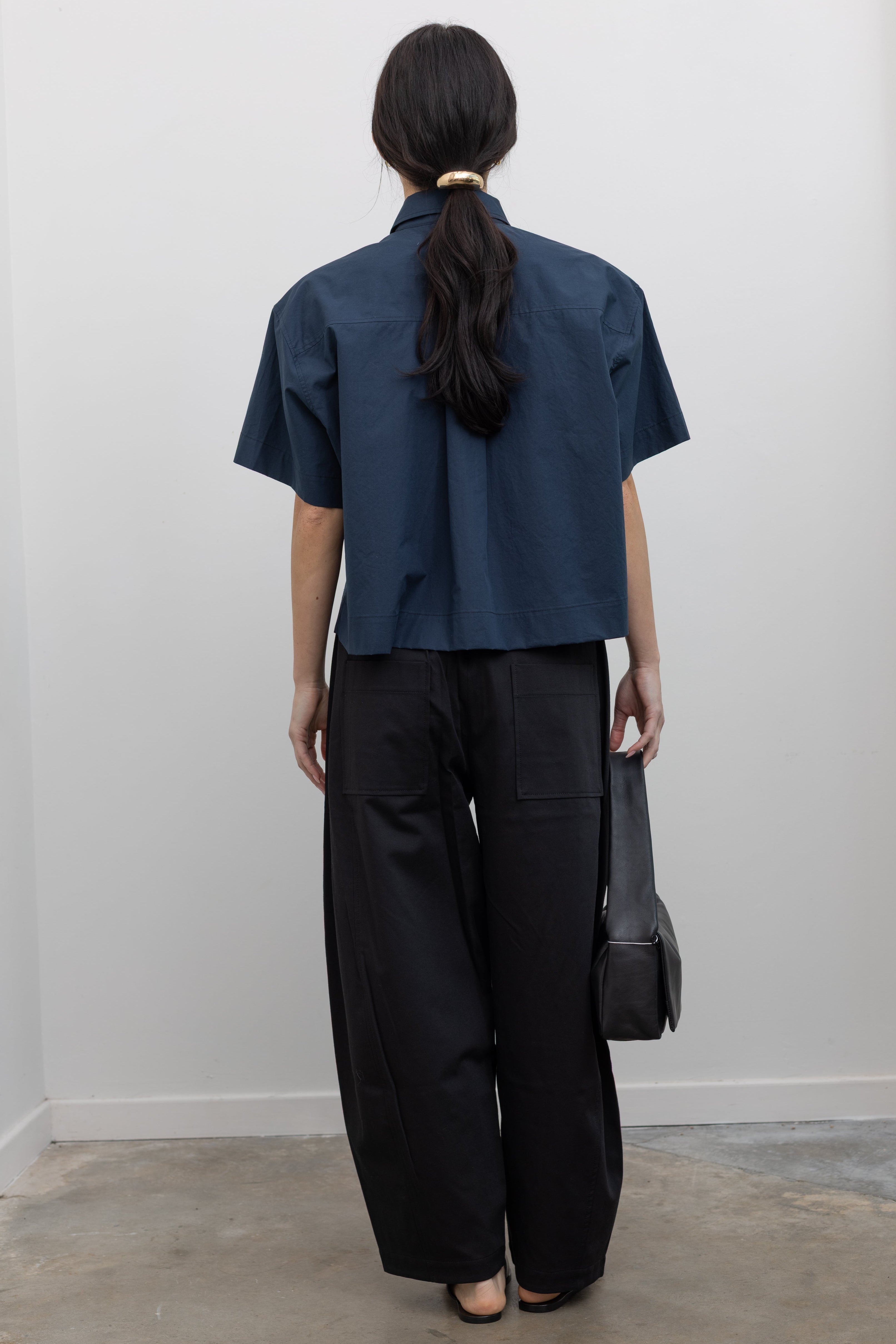 Full length back view of contemporary womenswear spring fashion in Mijeong Park black baggy balloon pants with curved outseams, a relaxed fit, elastic waistband, and back patch pockets paired with a structured navy blue short sleeve cropped hem button down shirt and a black leather St. Agni bag at an Alas Eius store. 