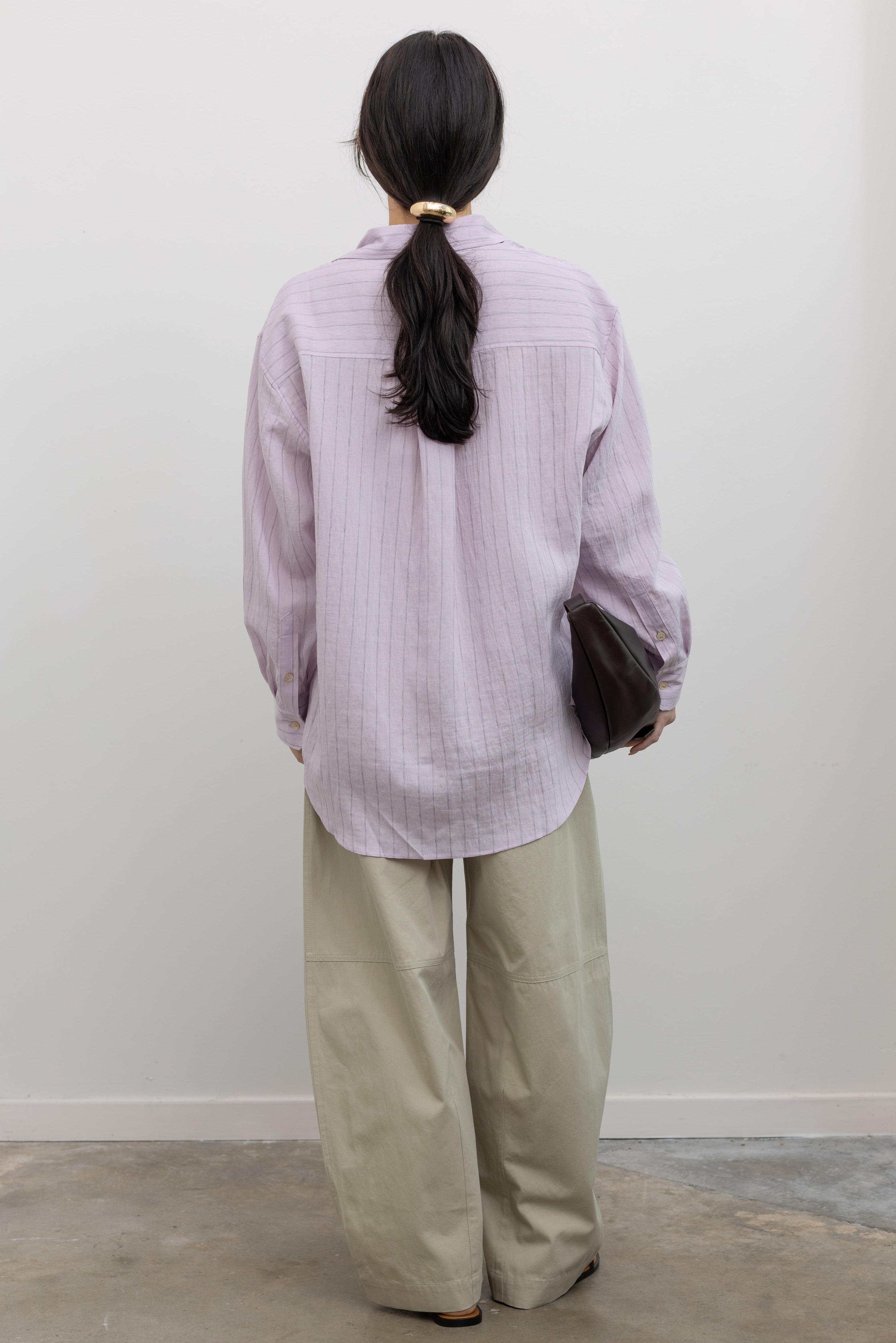 Full length back view of a woman wearing modern minimalist spring fashion in a Mijeong Park oversized soft pink pinstripe button down shirt featuring a classic collar, long sleeves, and an off center button down front paired with an elevated classic khaki baggy curved leg pant with strappy sandals at an Alas Eius store. 