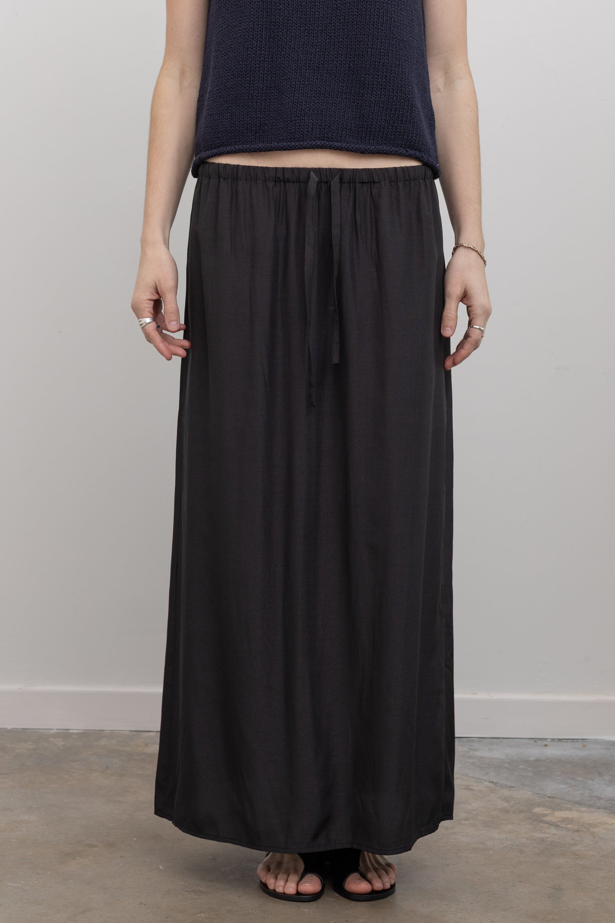 Close-up front view of a woman wearing contemporary spring fashion in a Mijeong Park black midi to maxi length skirt featuring an elastic drawstring waistband and discreet back slit paired with a Mijeong Park navy blue boat neck knit tank and strappy sandals at an Alas Eius store. 