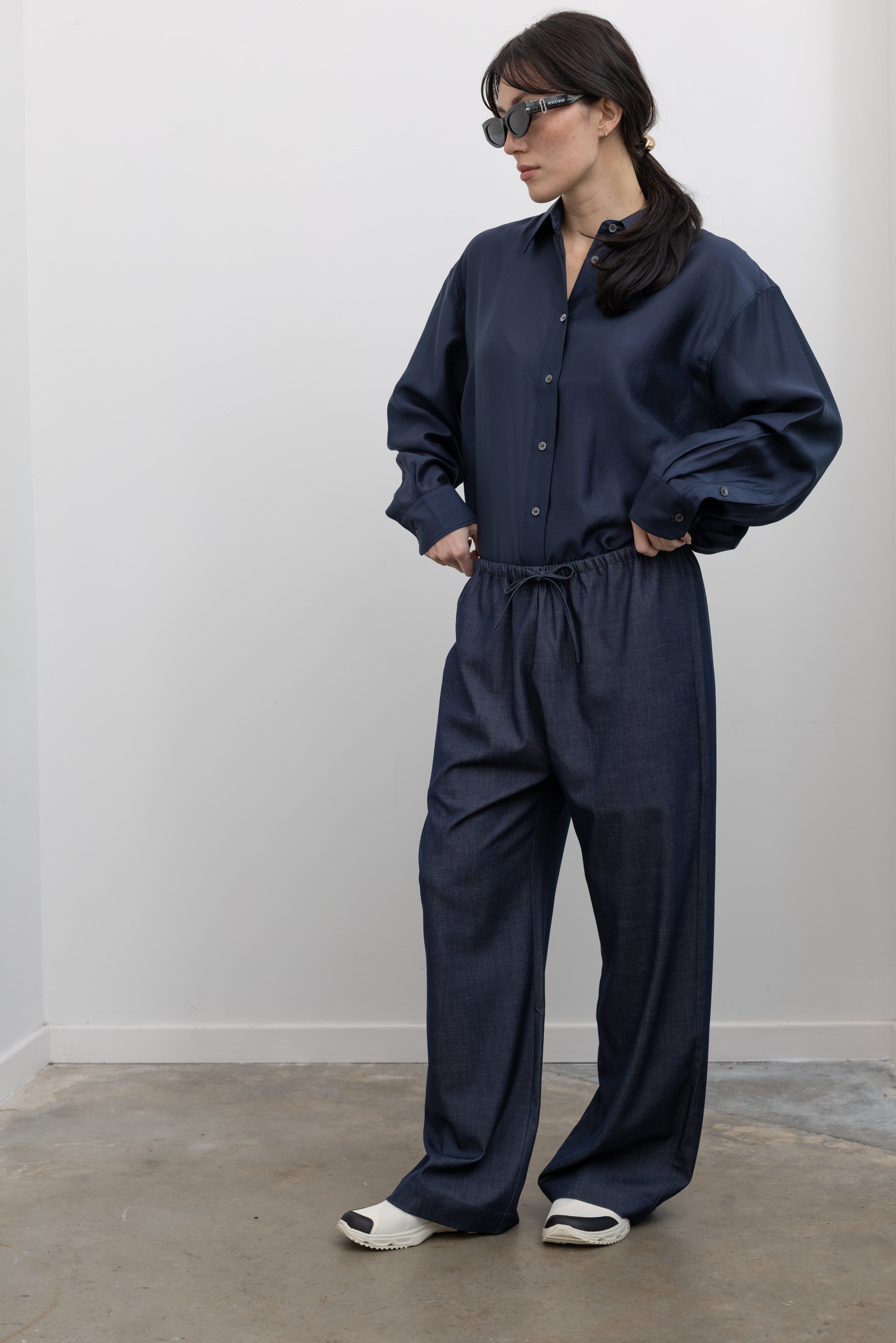 Full length 3/4 view of a woman standing wearing spring elevated essentials in Mijeong Park lightweight dark wash blue denim drawstring pants featuring an elastic waistband and side seam pockets paired with a navy blue semi sheer button down shirt and Salomon slide sneakers at an Alas Eius store. 
