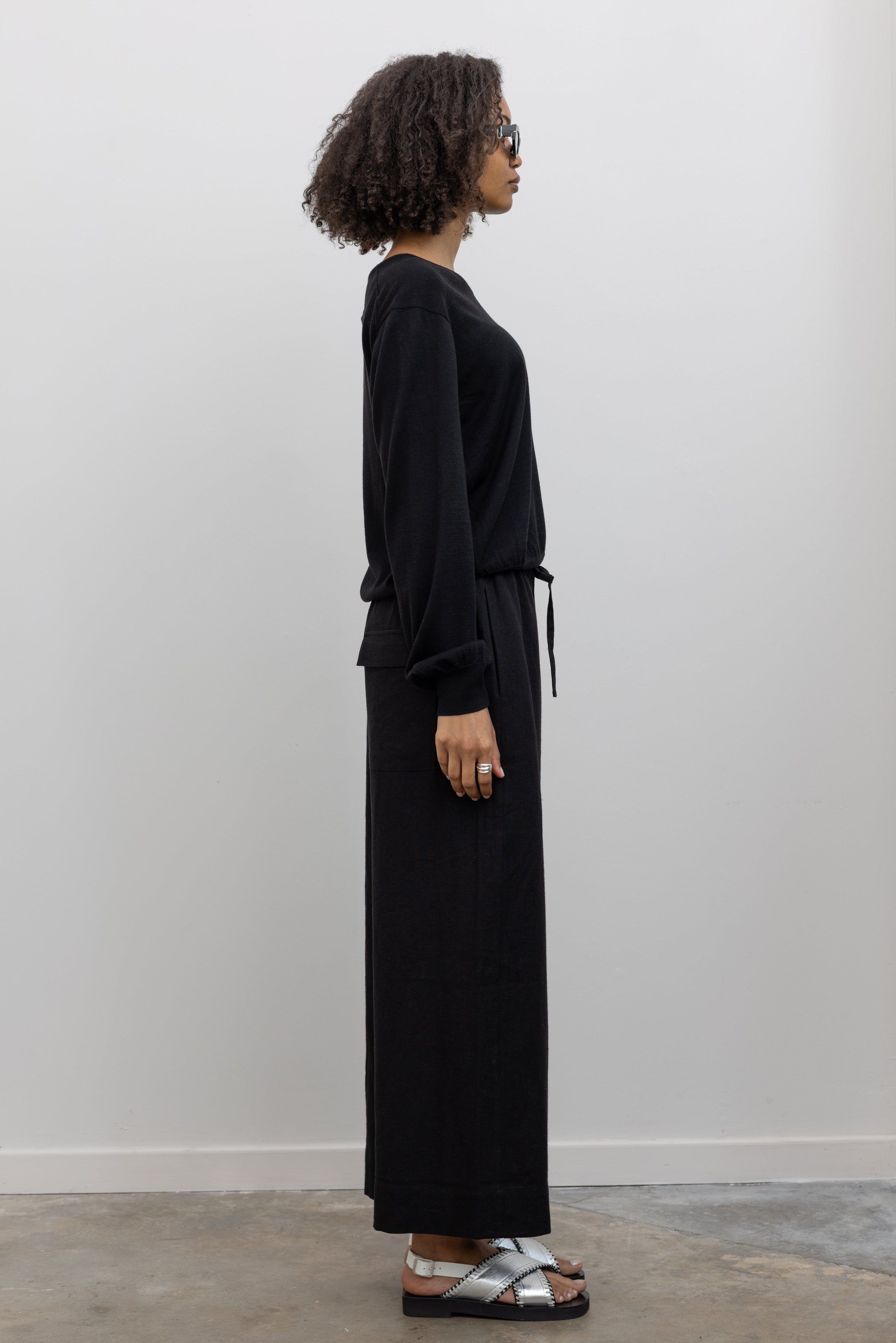 Full length side view of a woman standing wearing elevated casual weekend fashion in a Mijeong park Black Cotton Blend V-Neck Long Sleeve Knit Top paired with Mijeong Park Black Linen Blend Cropped Pants featuring a comfortable drawstring waist, front and back patch pockets, and a cropped length suitable for a minimalist, versatile wardrobe at an Alas Eius store.