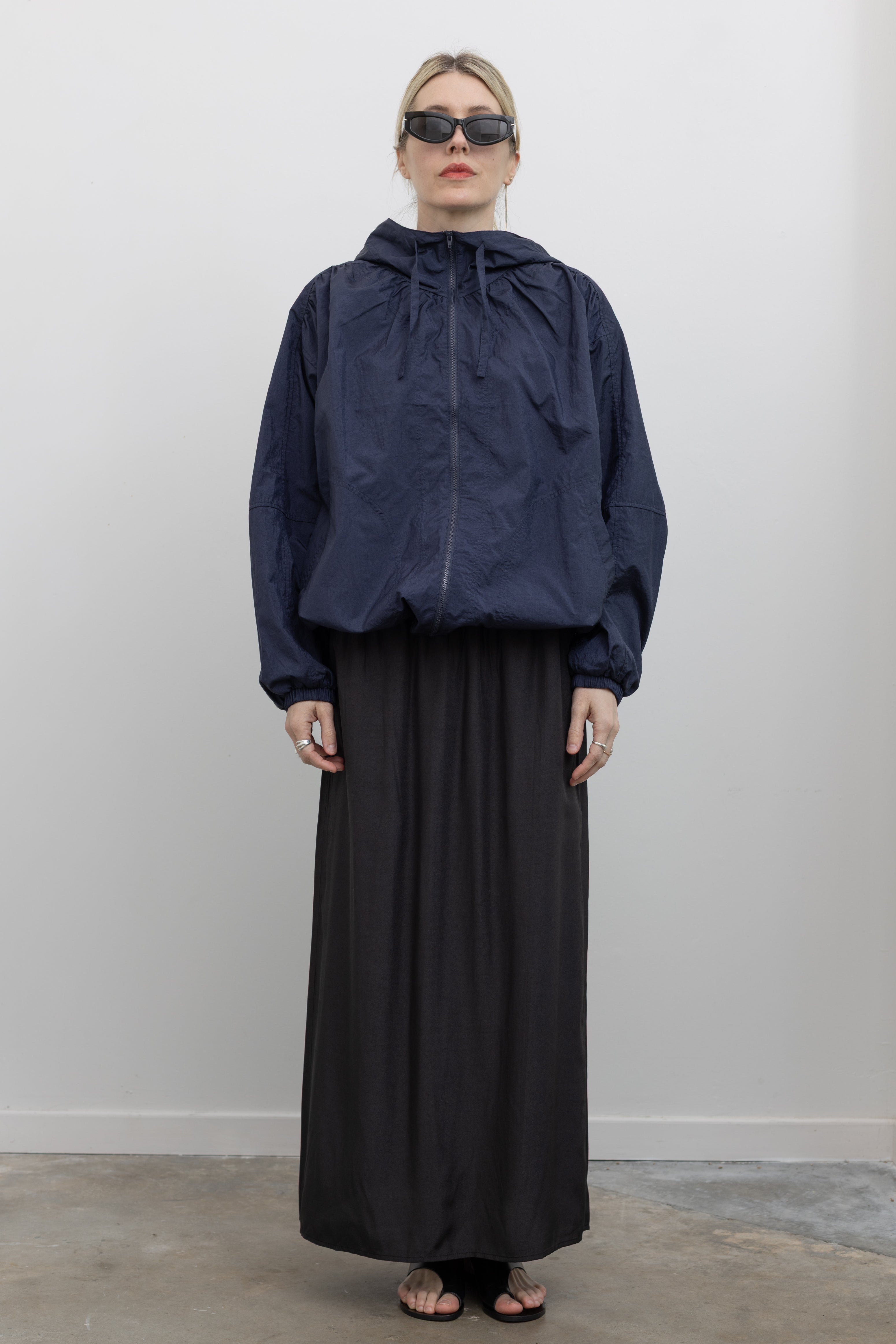 Full length front view of a woman wearing an elevated outerwear staple in a  Mijeong Park navy blue nylon shirred anorak jacket featuring an oversized fit, front pockets, hood, and a zipper closure at an Alas Eius store. 
