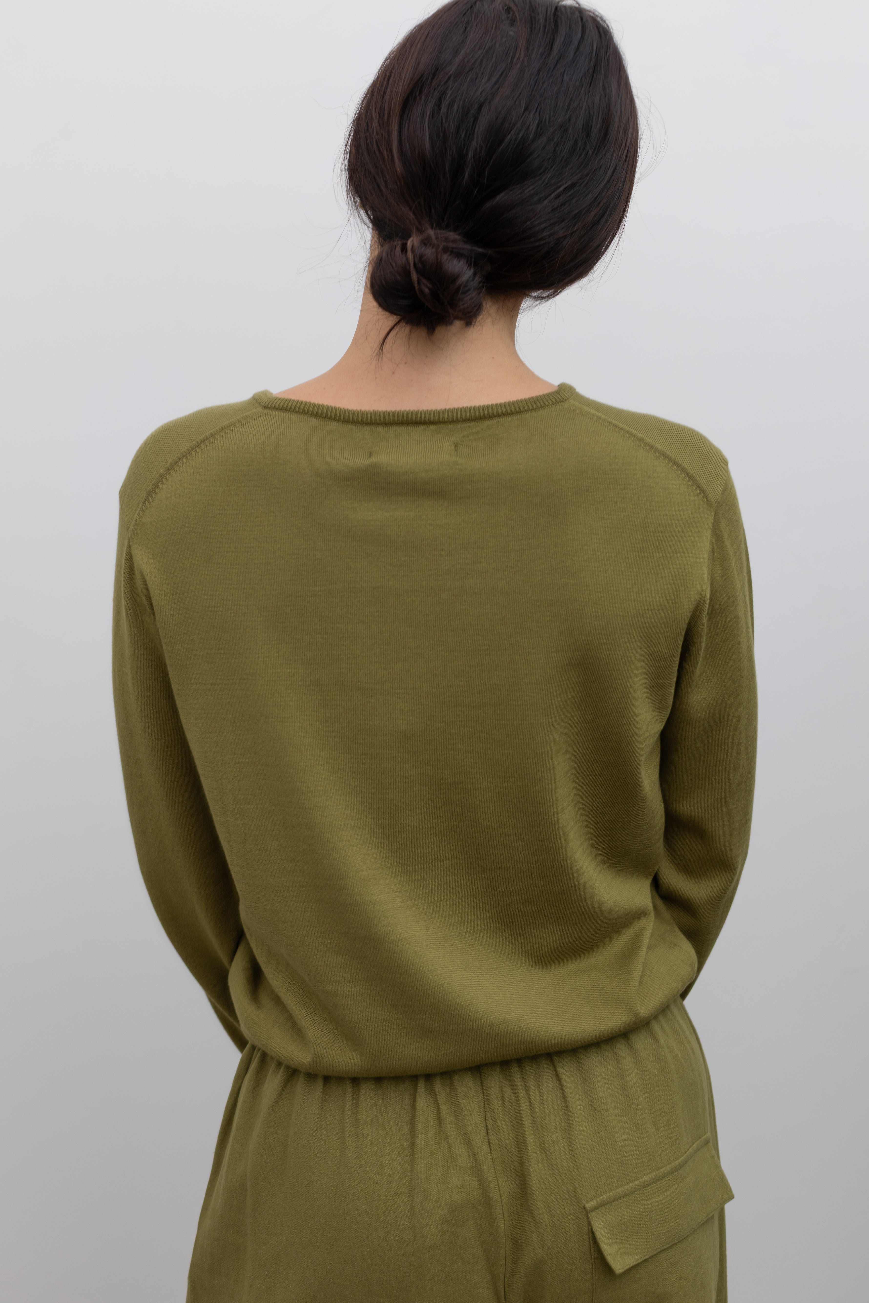 Back view of a woman posing wearing elevated capsule wardrobe essentials in the Mijeong Park Soft Green Cotton Blend V-Neck Knit Long Sleeve Top paired with Mijeong Park Green Linen Blend Wide Leg Cropped Pants featuring a comfortable drawstring waist, front and back patch pockets, and a cropped length suitable for a minimalist, versatile wardrobe at an Alas Eius store. 