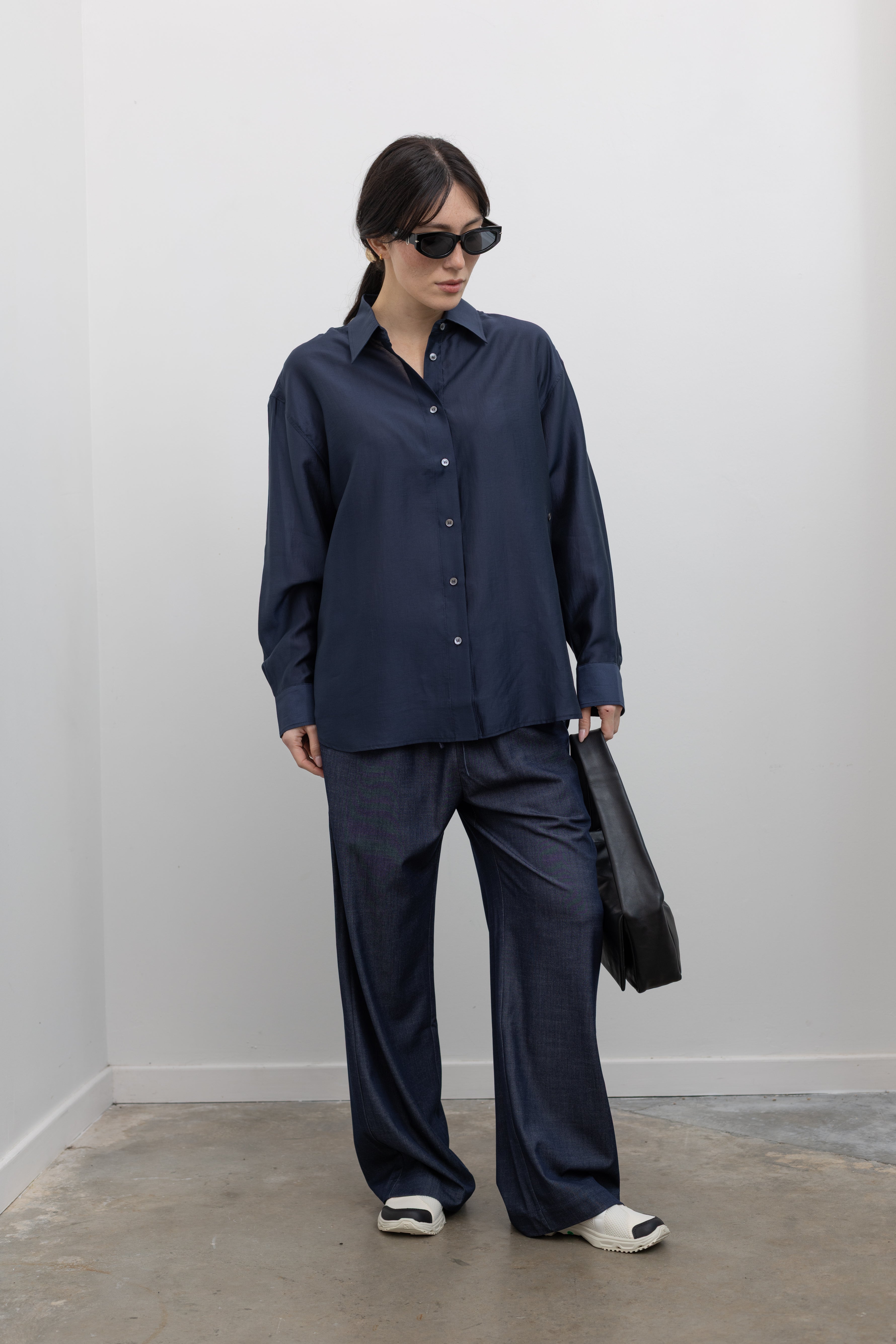 Full length front view of a woman wearing spring fashion in a Mijeong Park navy blue semi-sheer oversized shirt featuring a flattering off-center button front, long sleeves, and a classic sharp collar paired with Mijeong Park lightweight dark wash denim drawstring pants, Salomon slide sneakers, and a leather St. Agni bag at an Alas Eius store. 