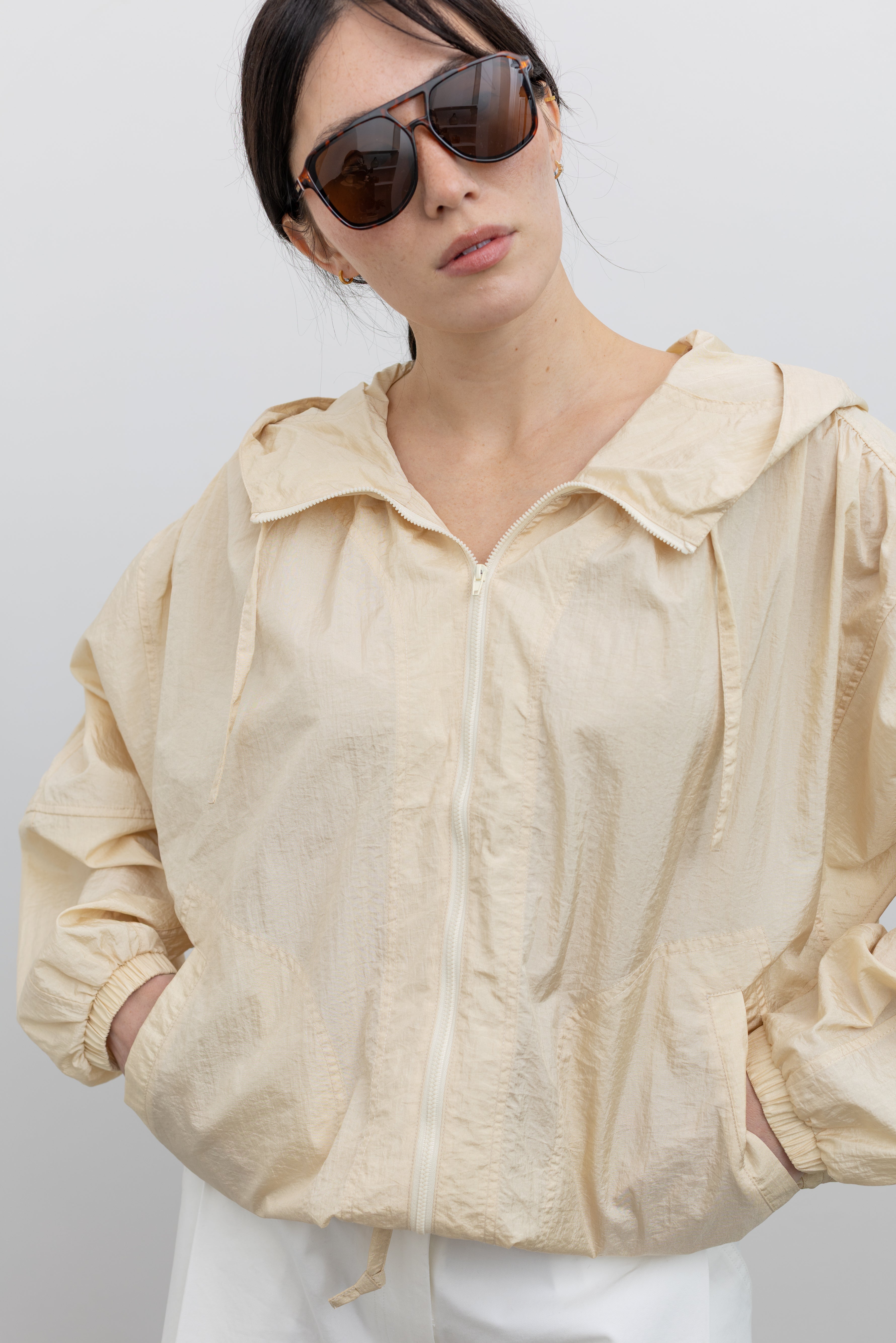 Close-up front view of a woman wearing an elevated outerwear staple in a Mijeong Park cream colored nylon shirred anorak jacket featuring an oversized fit, front pockets, hood, and a zipper closure at an Alas Eius store. 