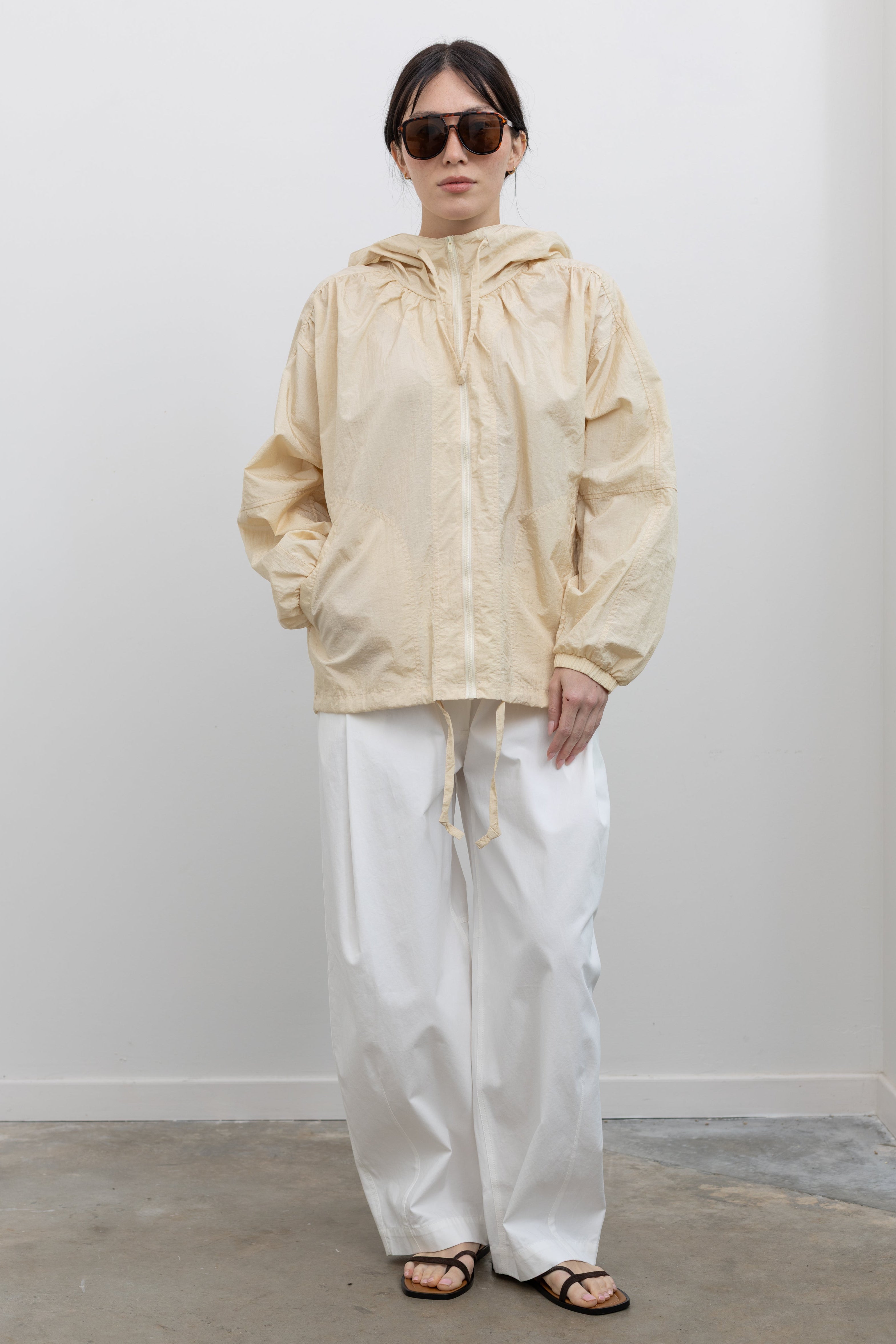 Full length front view of a woman wearing a versatile yet chic outerwear staple in a Mijeong Park cream colored nylon shirred anorak jacket featuring an oversized fit, front pockets, hood, and a zipper closure paired with white curved leg pants at an Alas Eius store. 
