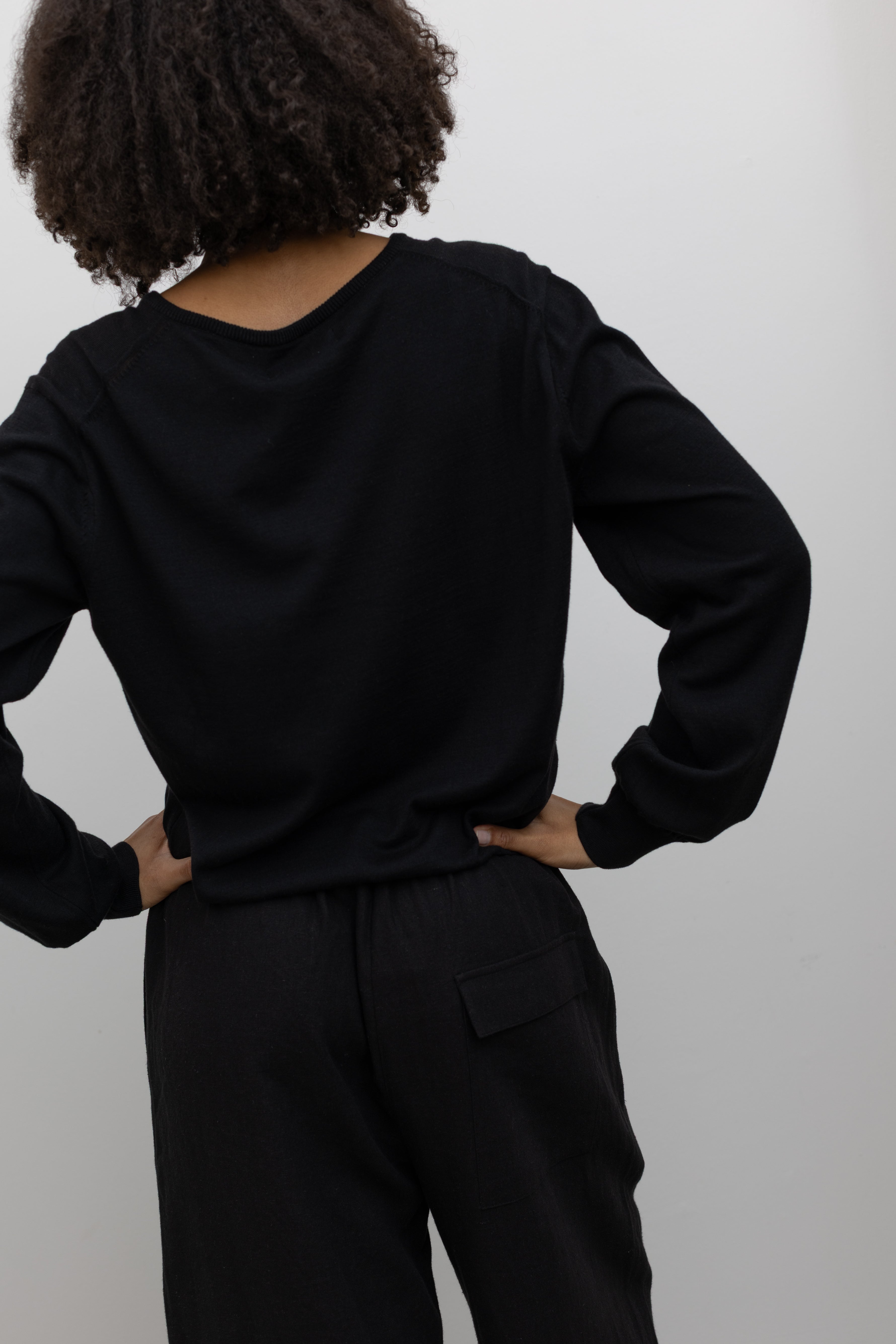 Close-up back view of a woman standing wearing elevated casual weekend fashion in a Mijeong park Black Cotton Blend V-Neck Long Sleeve Knit Top paired with Mijeong Park Black Linen Blend Cropped Pants featuring a comfortable drawstring waist, front and back patch pockets, and a cropped length suitable for a minimalist, versatile wardrobe at an Alas Eius store.