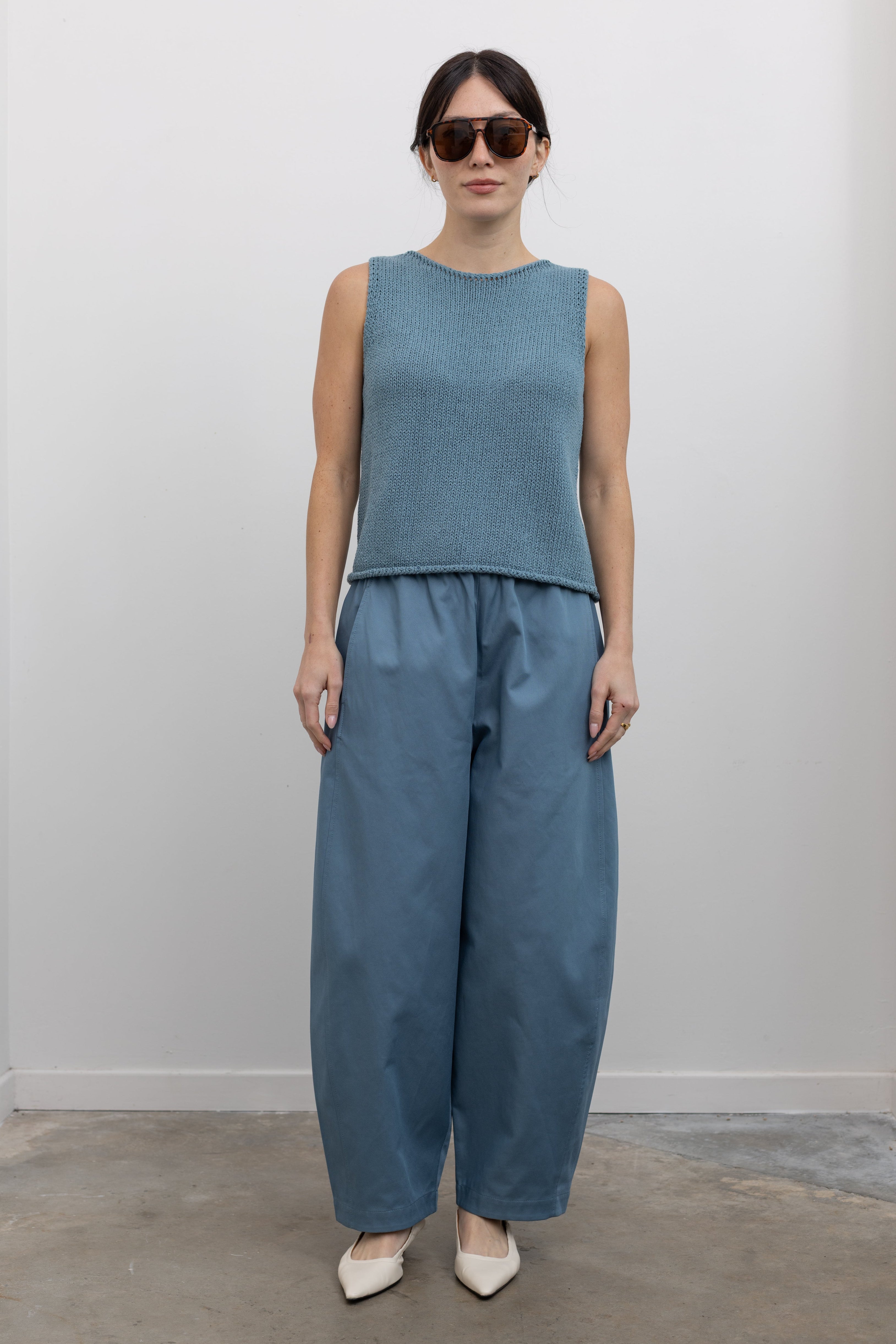 Full length front view of a woman wearing spring monochromatic fashion in Mijeong Park light blue cropped balloon pants with curved outseams, a relaxed fit, mid-rise waist, and a comfortable elasticated waistband paired with a Mijeong Park blue boat neck knit tank with white leather kitten heels at an Alas Eius store. 