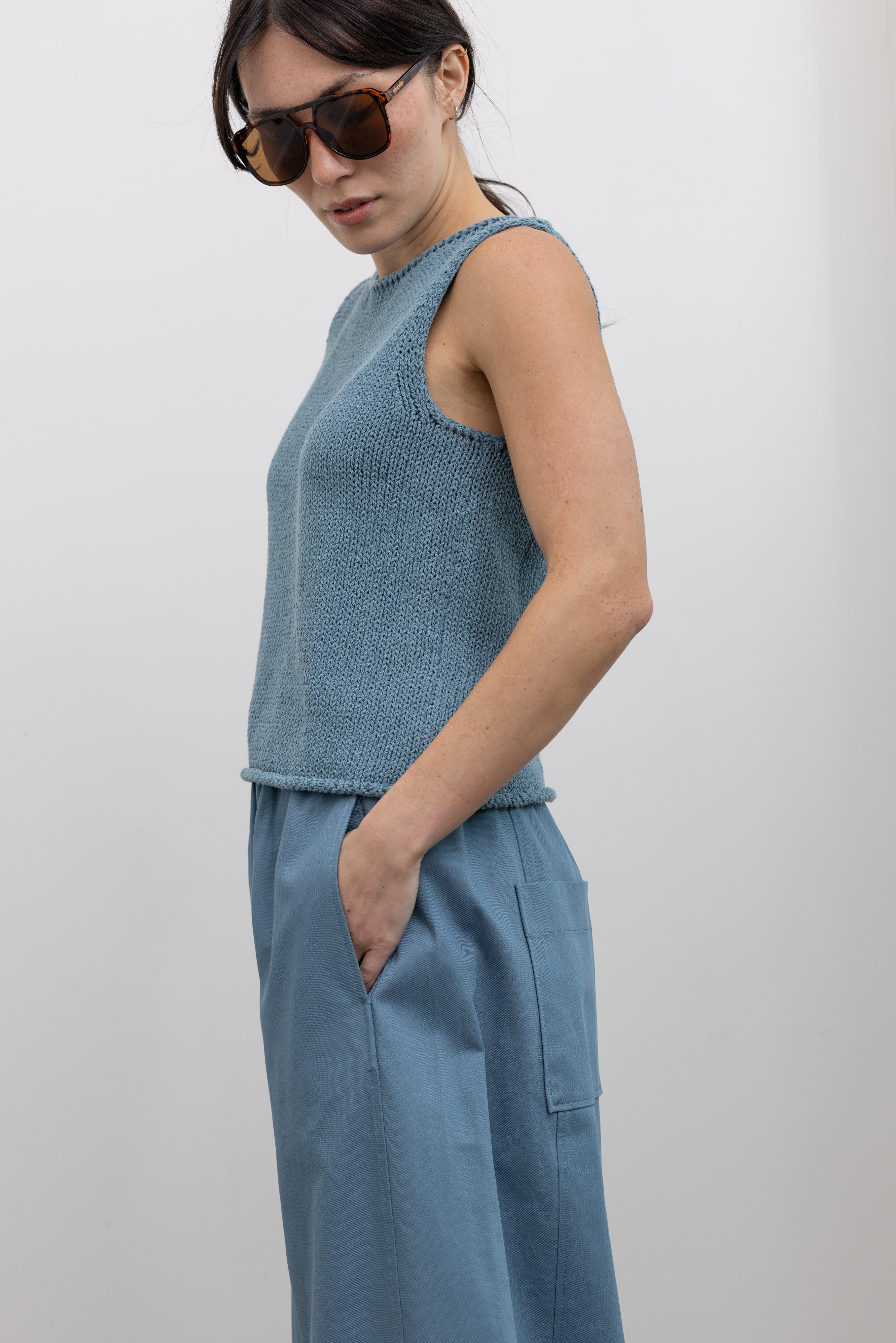 Close-up side view of a woman wearing spring fashion in a Mijeong Park light blue boat neck knit sweater tank top featuring rolled edges around the neckline and hem at an Alas Eius store. 