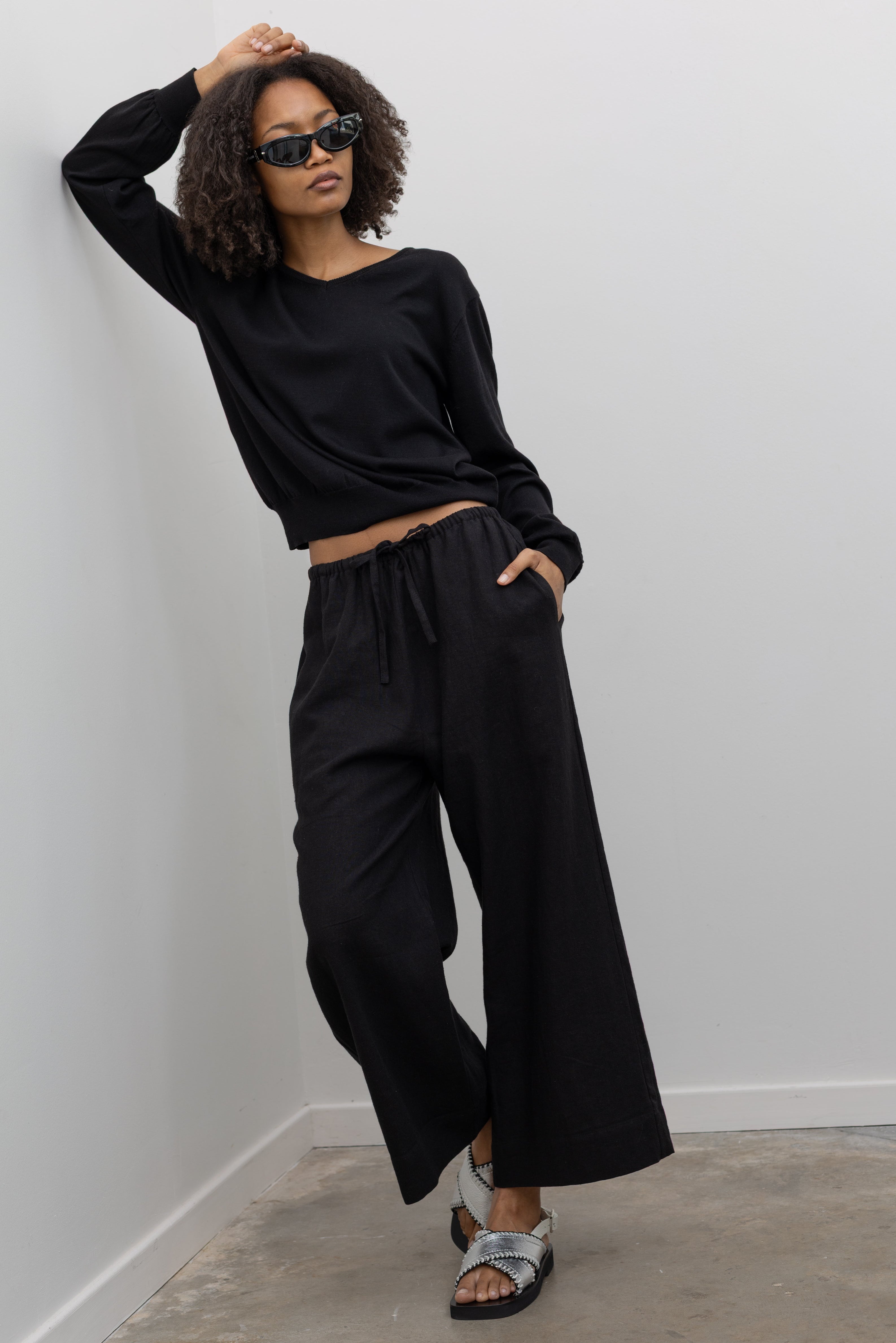 Full length front view of a woman posing wearing elevated casual weekend fashion in a Mijeong park Black Cotton Blend V-Neck Long Sleeve Knit Top paired with Mijeong Park Black Linen Blend Cropped Pants featuring a comfortable drawstring waist, front and back patch pockets, and a cropped length suitable for a minimalist, versatile wardrobe at an Alas Eius store.